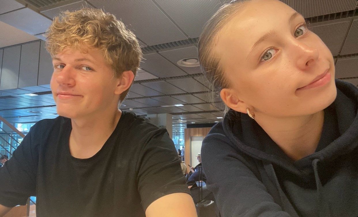 A young boy with curly blonde hair and a young girl with blonde hair pulled back are smiling at the camera.