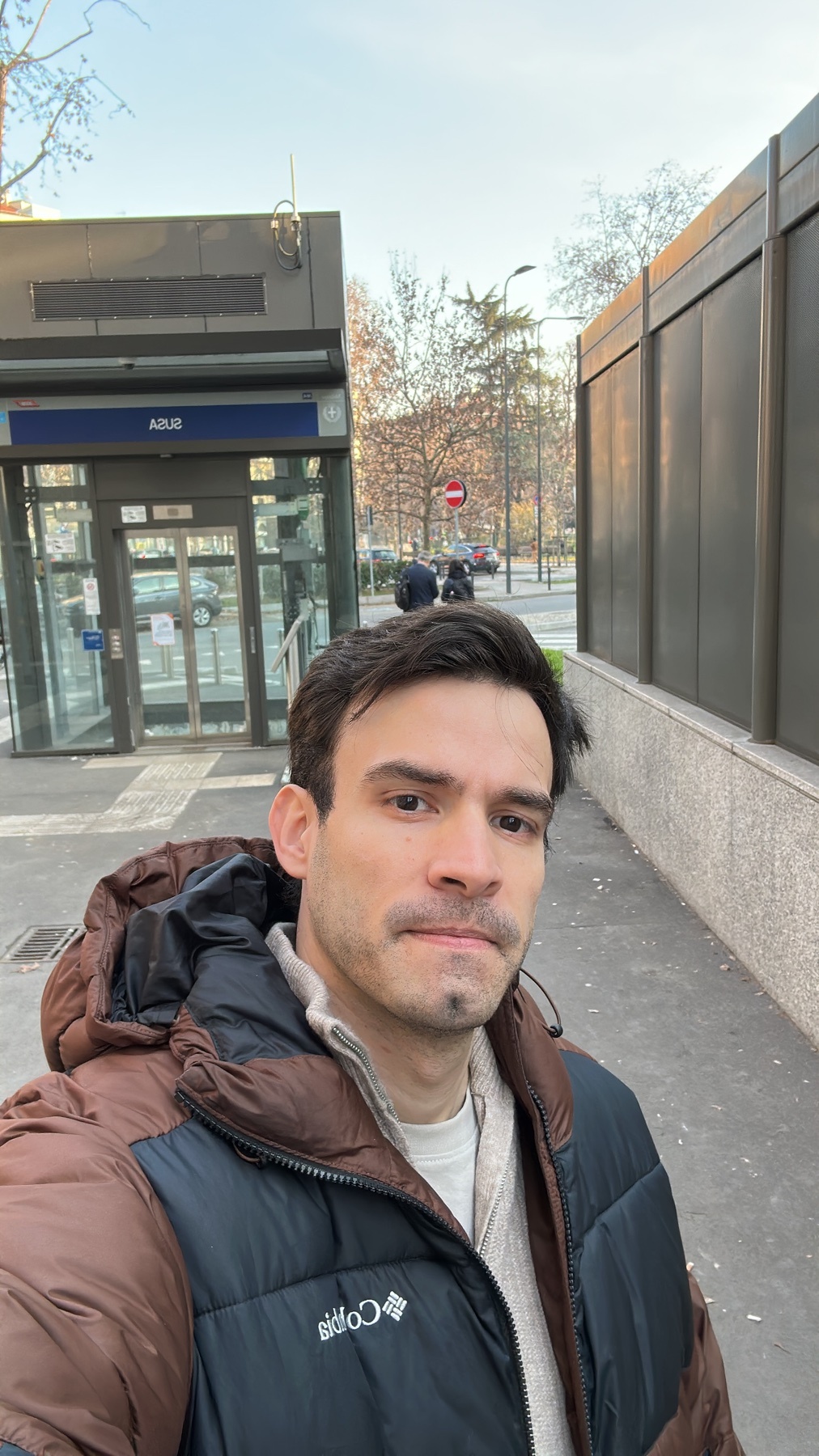 A man takes a selfie in front of a modern building with glass doors. He is wearing a brown and black puffy jacket and looking towards the camera.