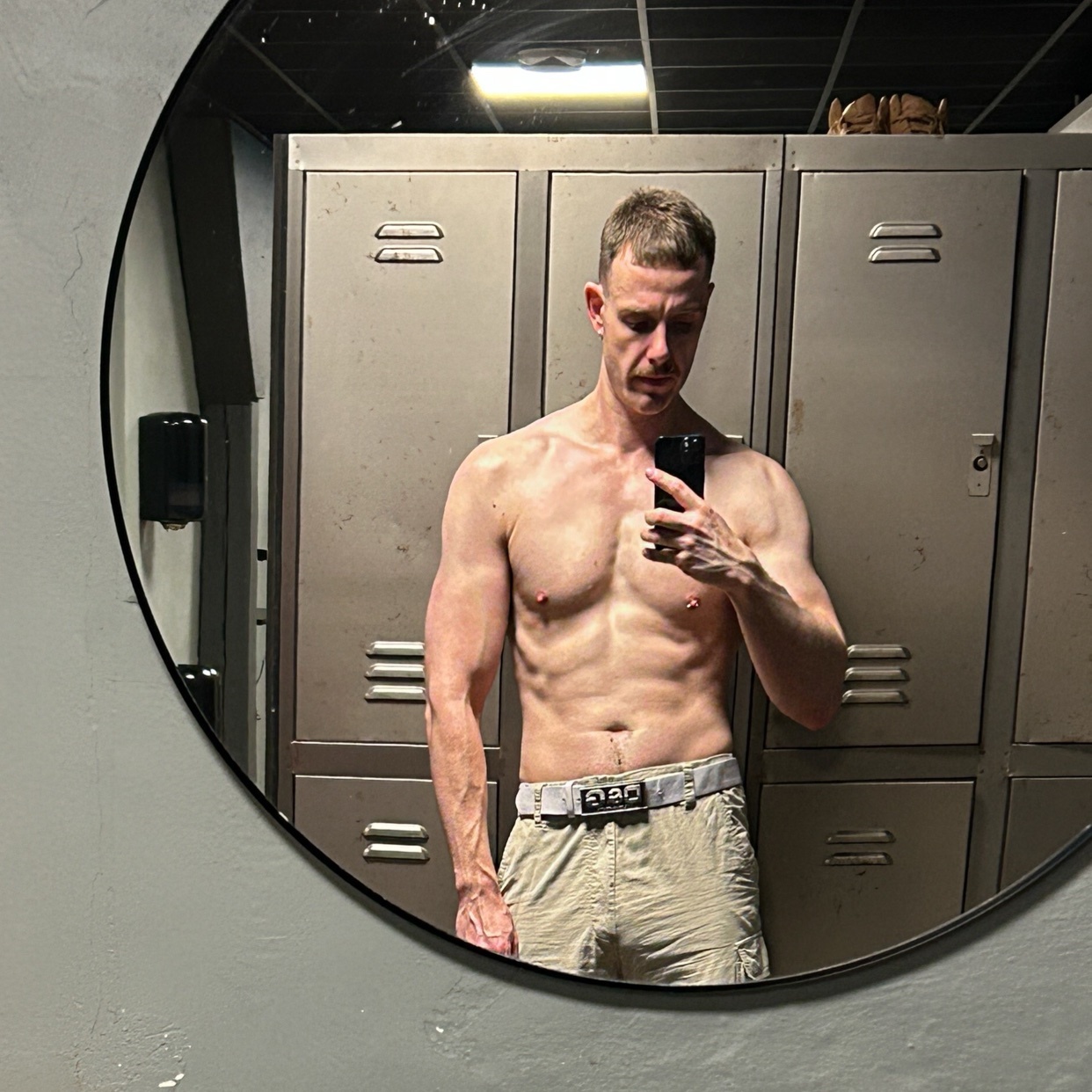 A man with a mustache and stubble takes a mirror selfie in a locker room, showing off his muscular physique in gym shorts.