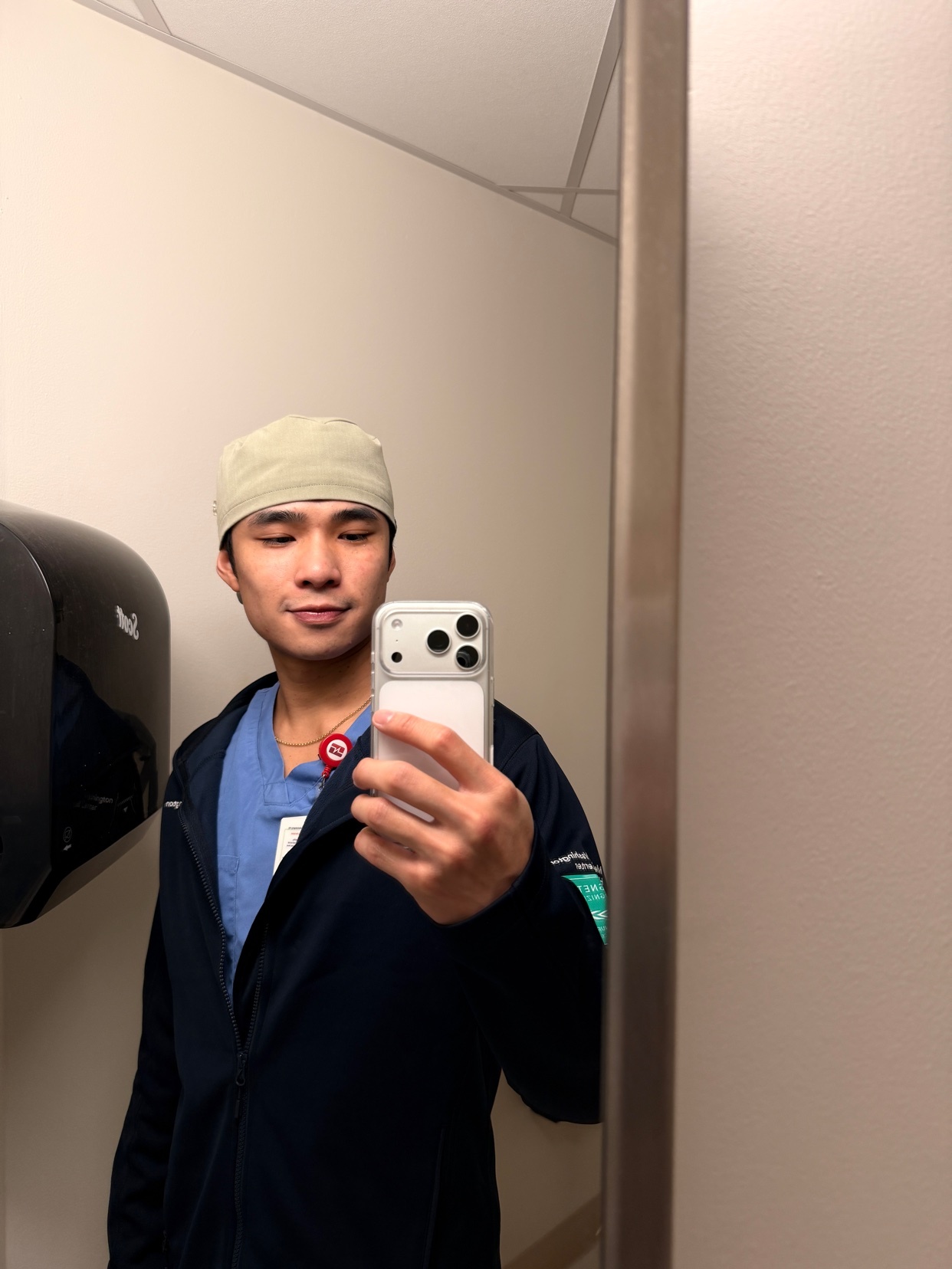 A man takes a mirror selfie while wearing scrubs and a cap. The photo appears to be taken in a restroom.