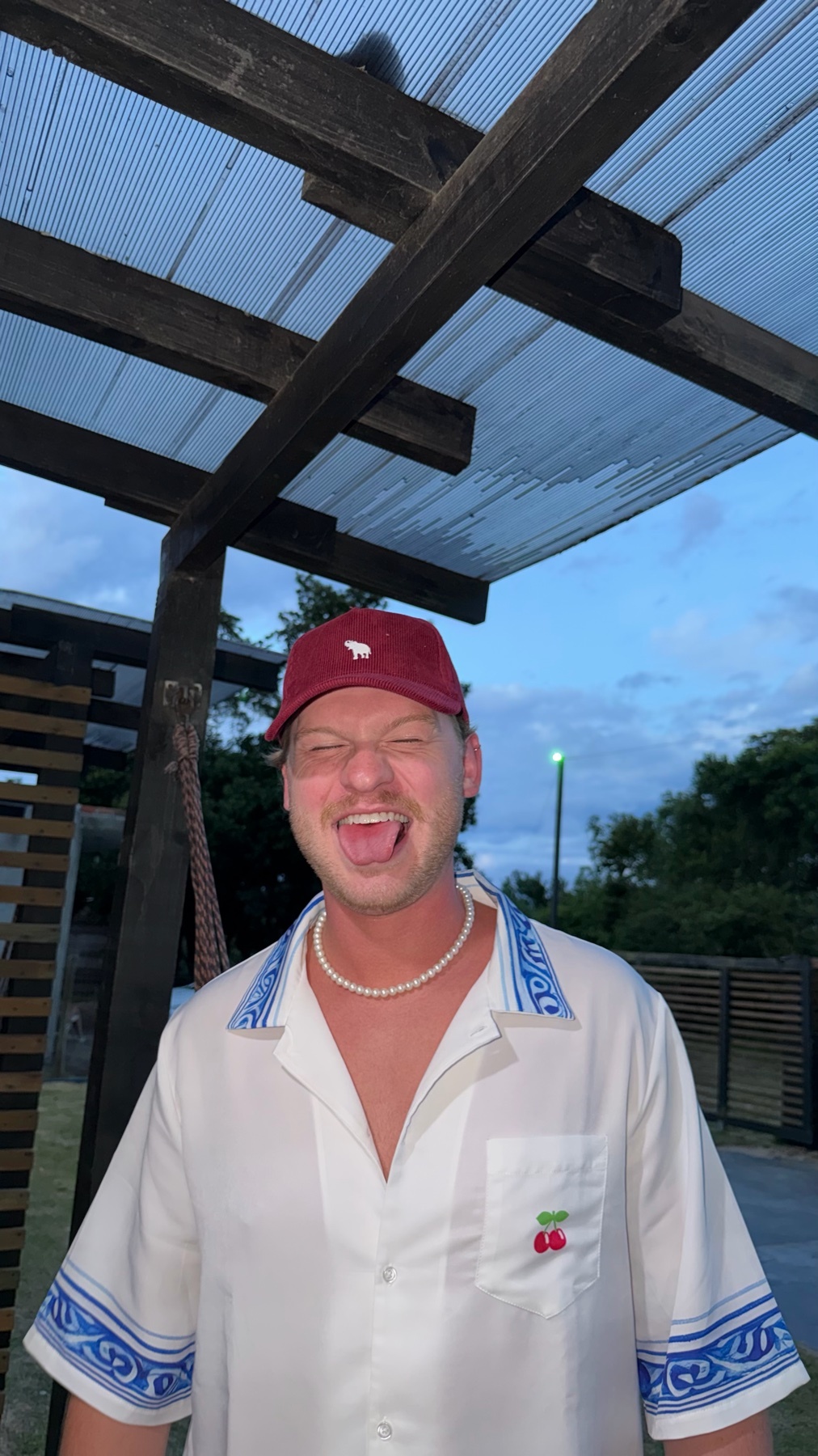 A man is smiling and wearing a baseball cap and a pearl necklace. He is shirtless and wearing a white shirt with blue accents.