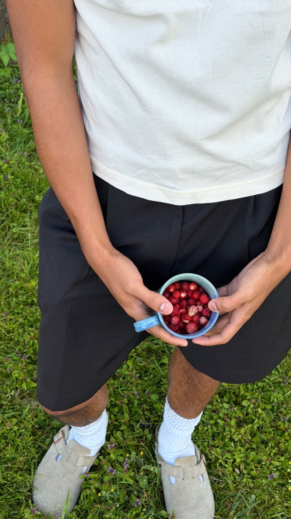 A person holds a small blue cup filled with wild strawberries. The person is wearing a white t-shirt, black shorts, white socks, and clogs. The background is green grass.