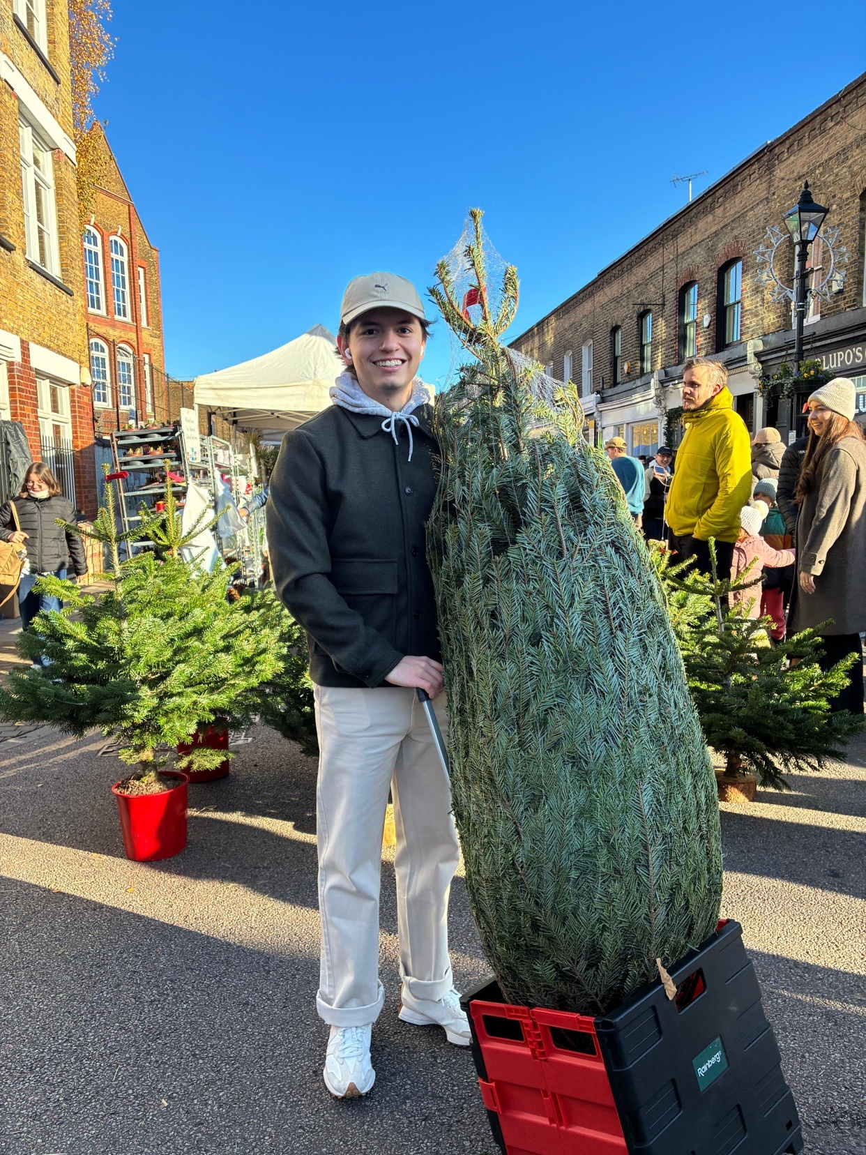 A man smiles while holding a large Christmas tree in a street market. The photo is taken during the day, with other trees and people in the background.