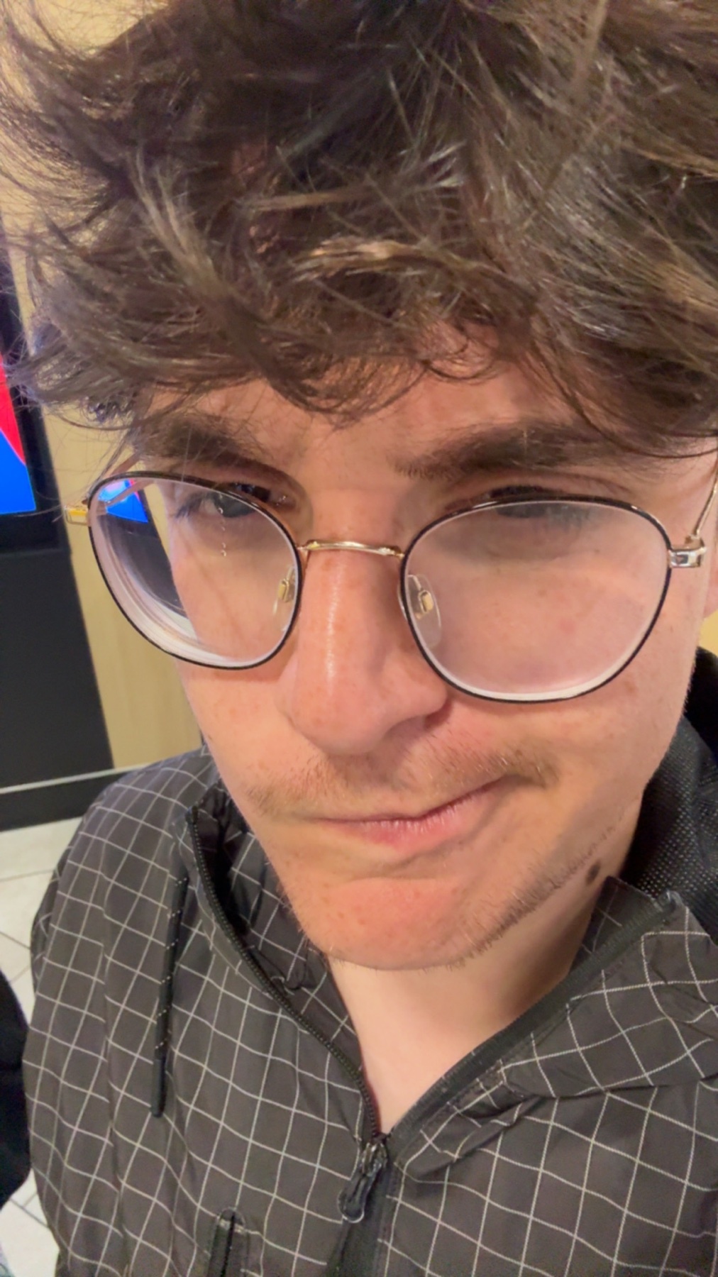 A close-up selfie of a young man wearing glasses and a patterned jacket. He has dark, messy hair and stubble on his face.