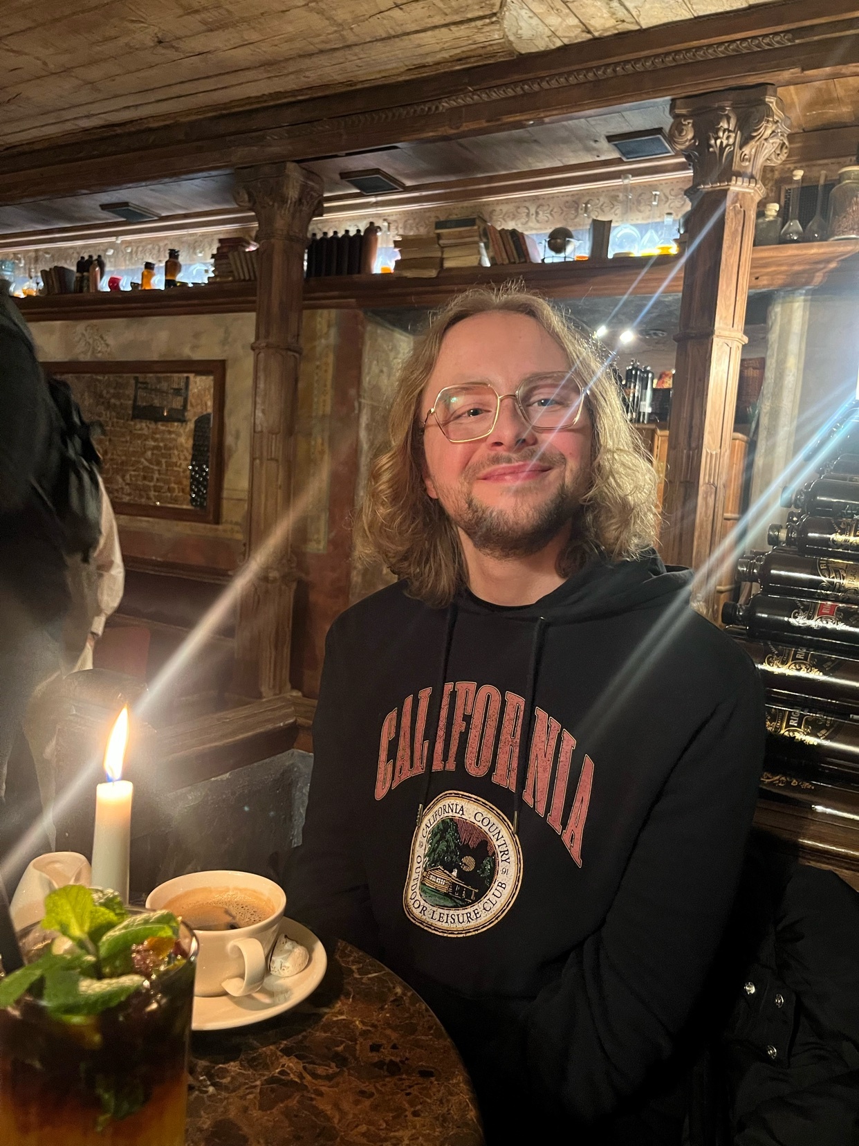 A man with blonde, curly hair and glasses smiles at the camera while sitting at a table in a restaurant. A candle and a drink are on the table.