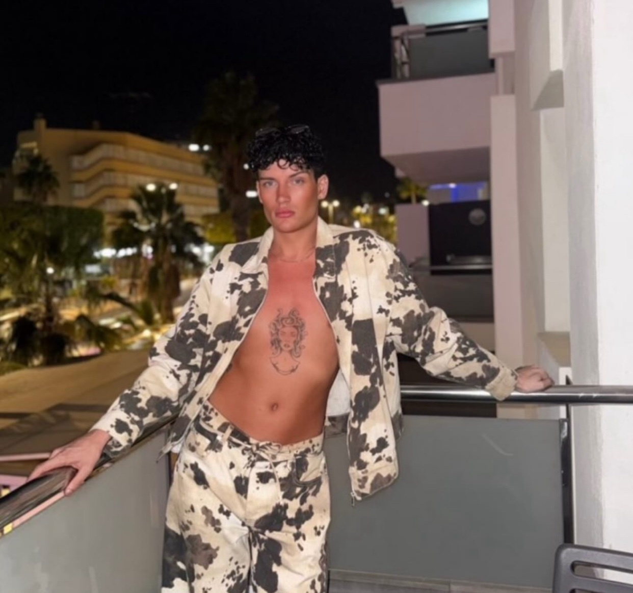 A man poses on a balcony at night, shirtless and wearing a cow-print jacket and pants. The photo is taken in Gran Canaria.