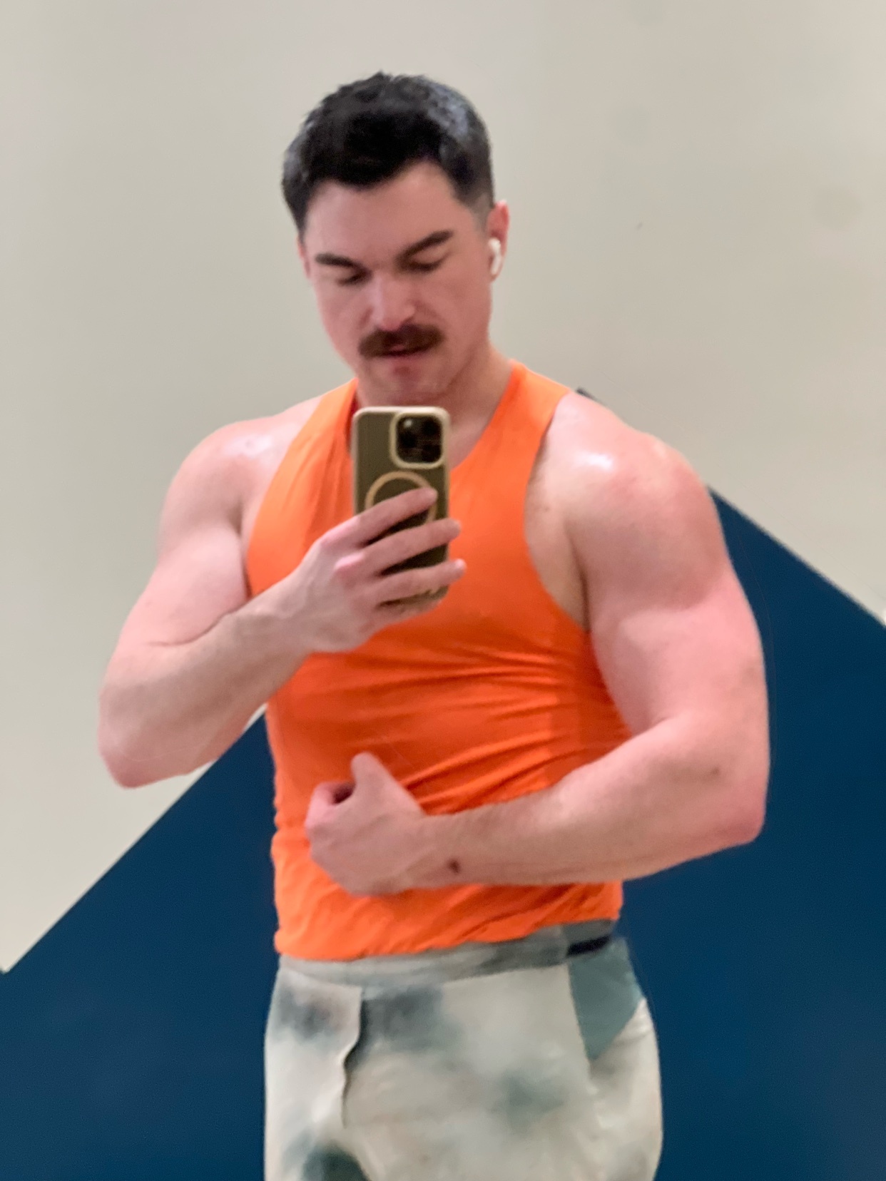 A man with a mustache takes a selfie in a gym, wearing an orange tank top and flexing his bicep. He appears sweaty, indicating a recent workout.