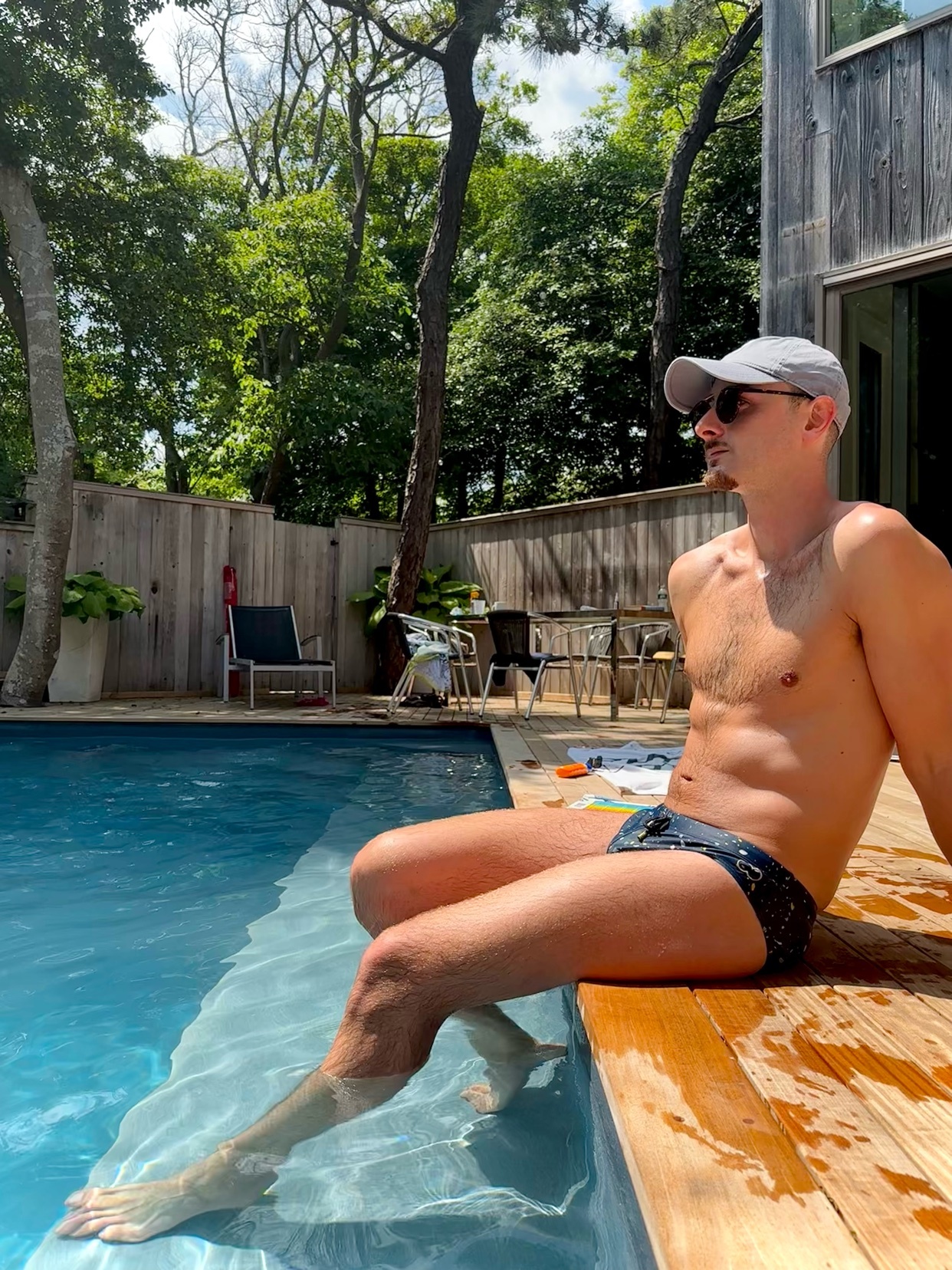 A man sits poolside in swim trunks, wearing sunglasses and a baseball cap. He is shirtless and appears to be enjoying a sunny day.