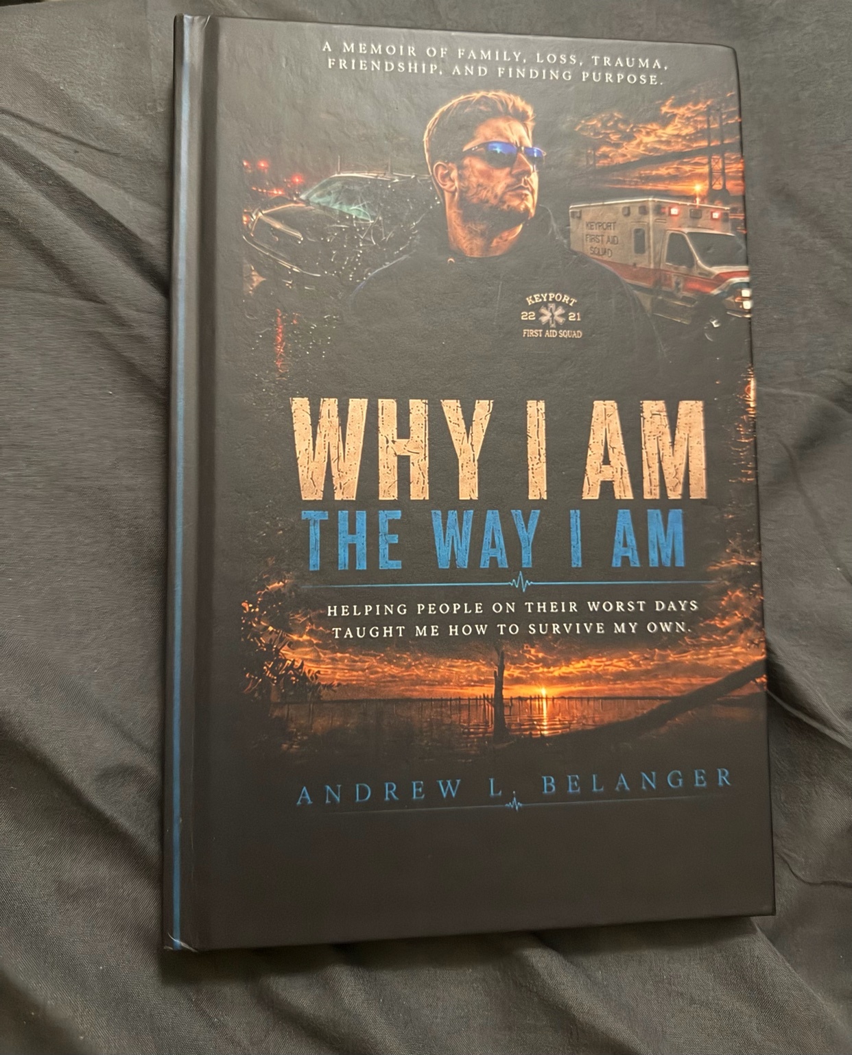 The cover of a memoir titled 'Why I Am The Way I Am' by Andrew L. Belanger. The cover features a man in sunglasses looking off into the distance, with an ambulance and a car in the background. A sunset is visible on the horizon.