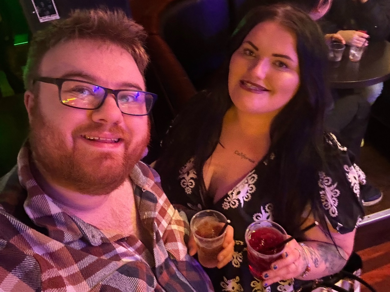 A selfie of a man and a woman smiling and holding drinks. They appear to be at a bar or club.
