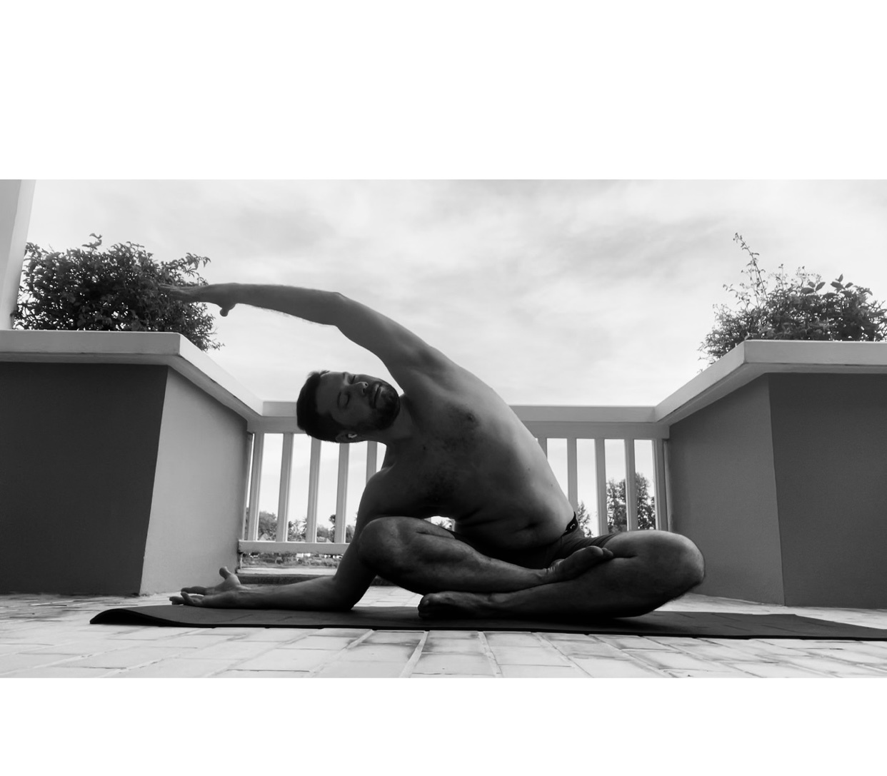 A man is doing yoga on a mat outdoors. He is shirtless and in a seated position, stretching his arm overhead.