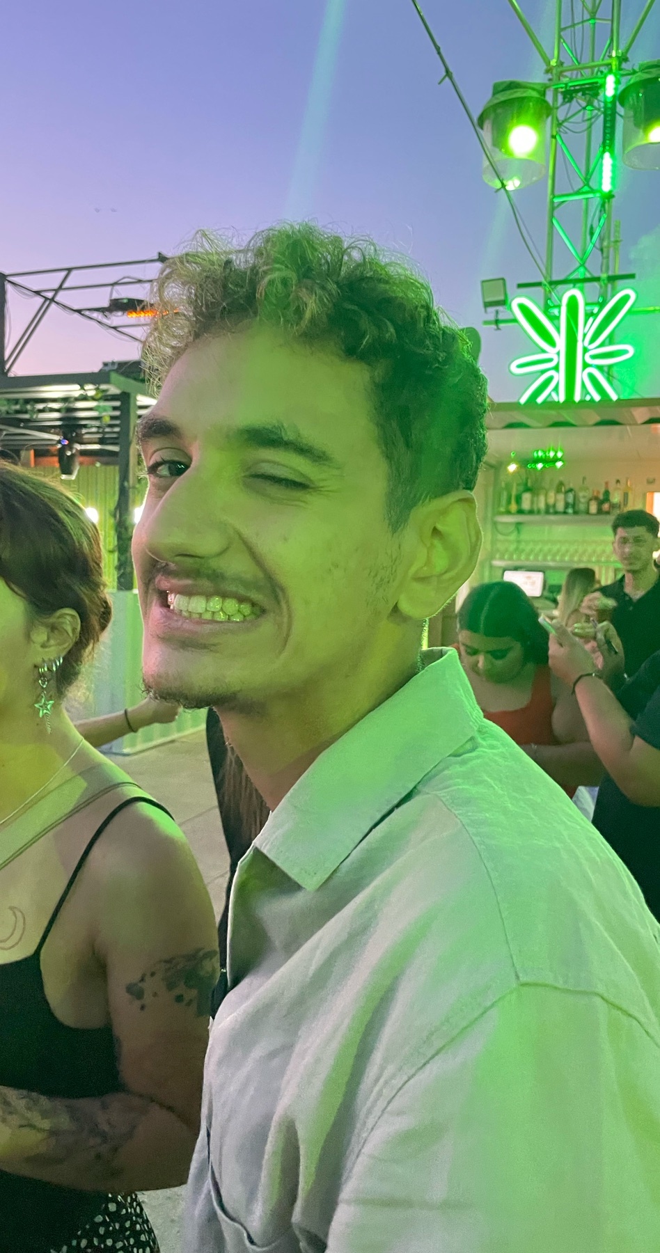 A man winks at the camera while at a party. He is smiling and surrounded by other people.