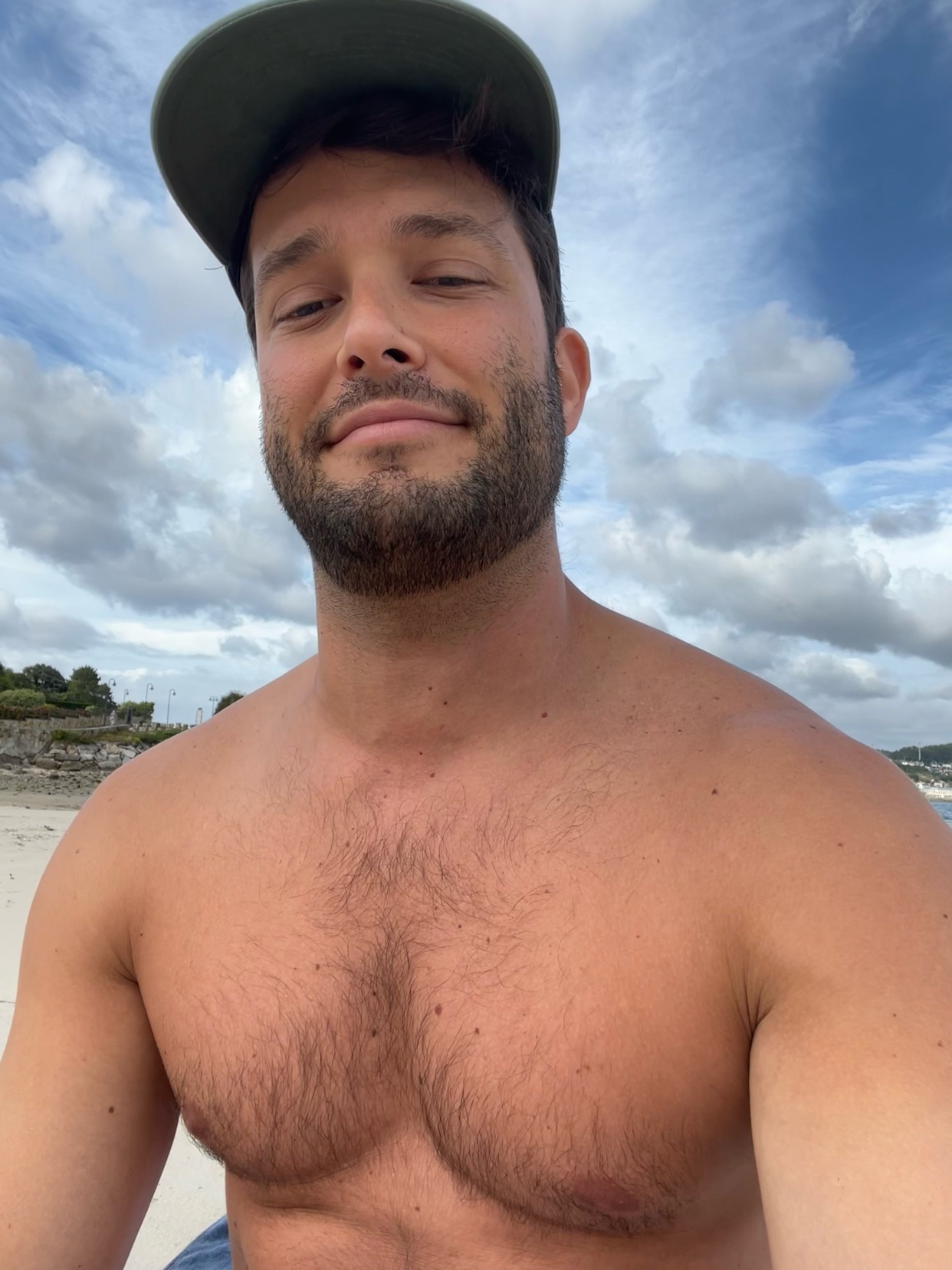 A man with stubble and chest hair smiles at the camera while wearing a baseball cap backwards. He is shirtless at the beach with a blue sky and clouds overhead.