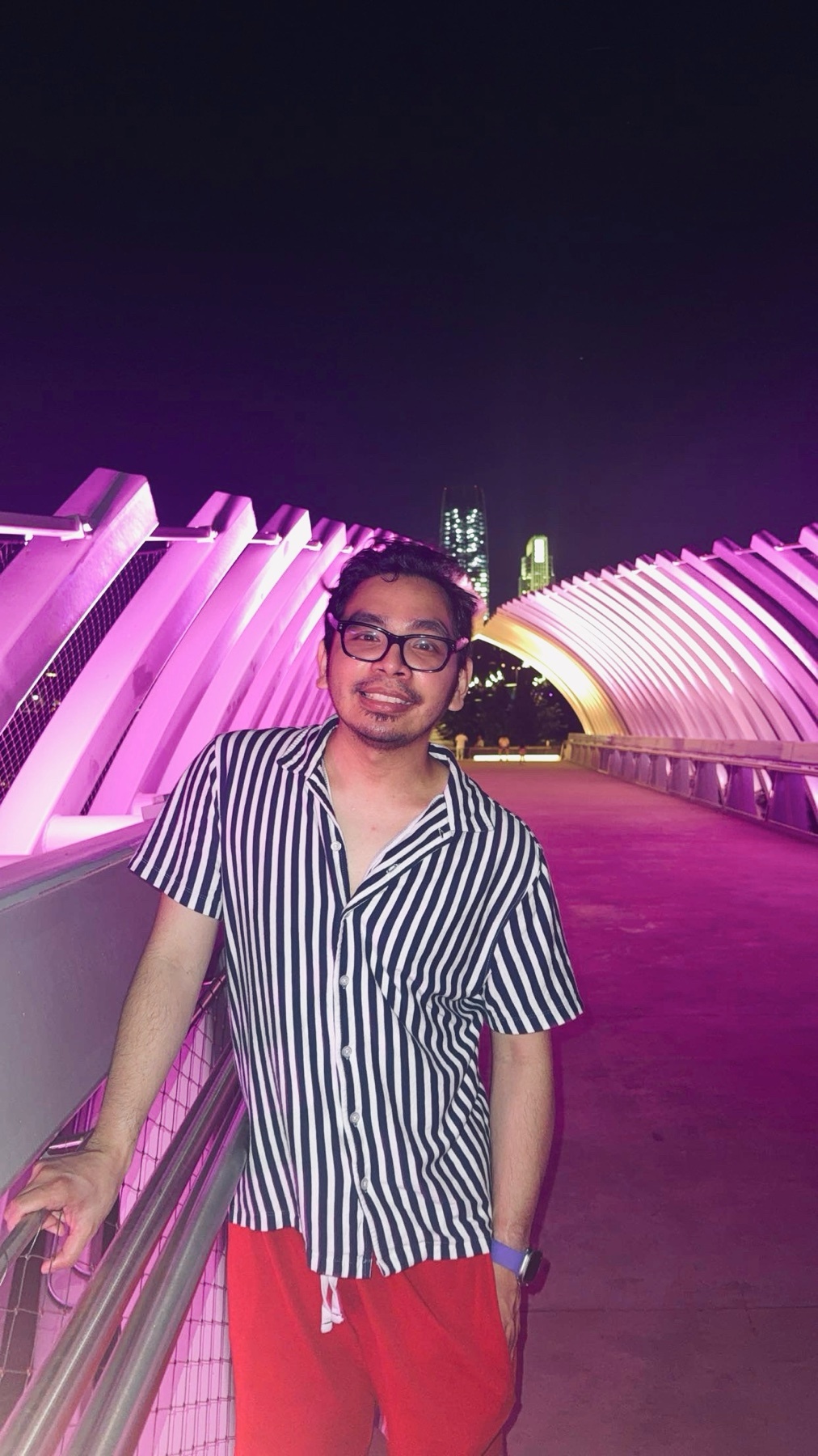 A man wearing glasses and a striped shirt poses on a bridge at night. The bridge is illuminated with pink and purple lights, with a cityscape visible in the background.