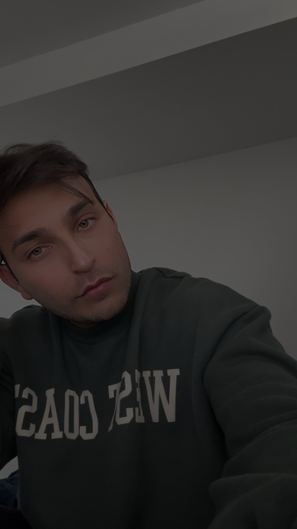 A man poses for a selfie, looking directly at the camera. He is wearing a dark green sweater with white text.