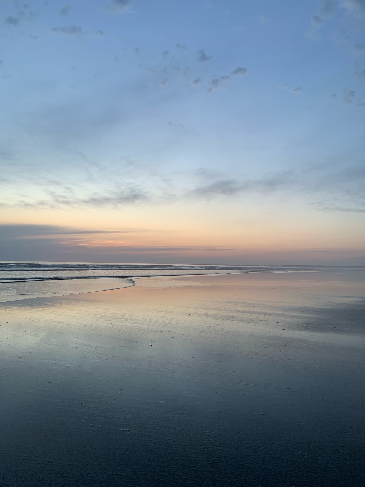 A serene beach scene at sunset or sunrise, with the sky reflecting on the wet sand. The pastel colors of the sky blend into the calm ocean.