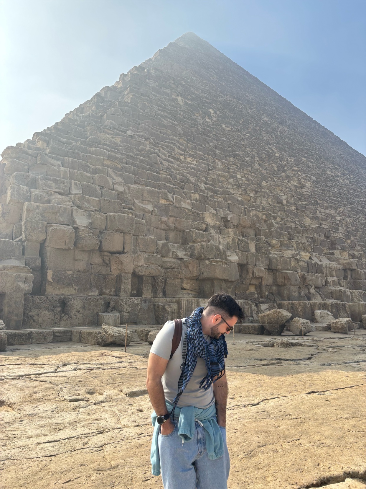 A man stands in front of a large pyramid in Egypt. He is wearing sunglasses and a scarf.