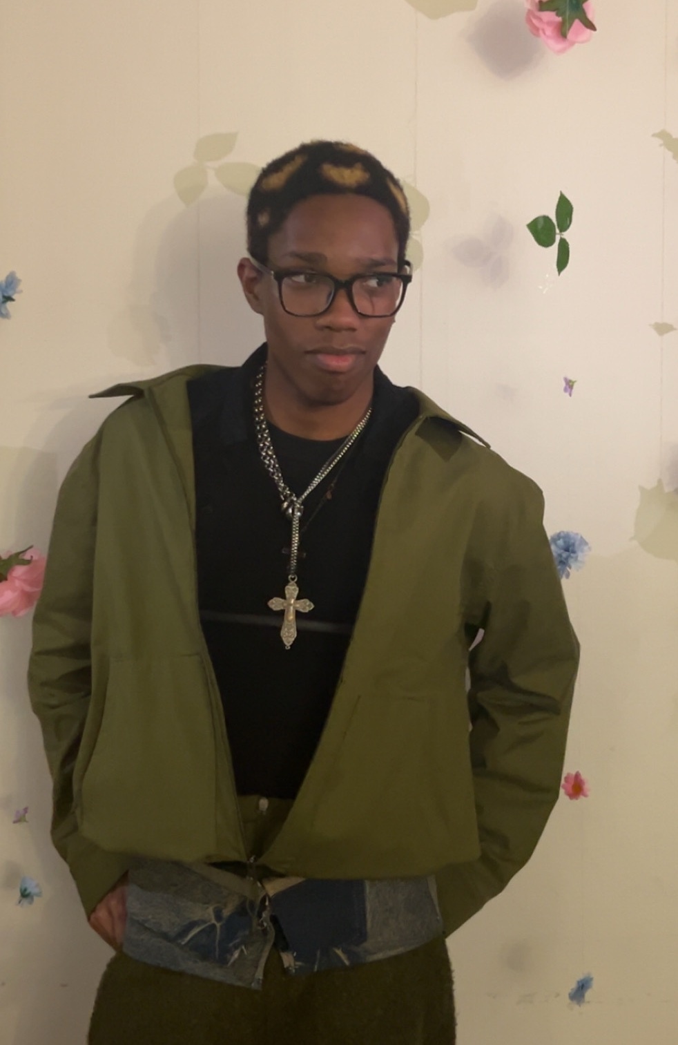 A man poses in a green jacket and black shirt, accessorized with a chain and glasses. The background features a floral wallpaper.