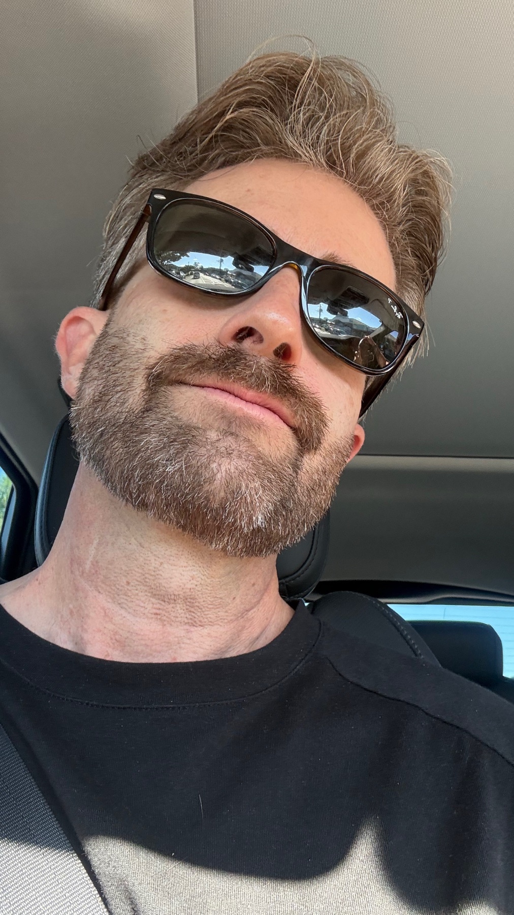 A man wearing sunglasses takes a selfie in a car. He has a beard and is wearing a black t-shirt.