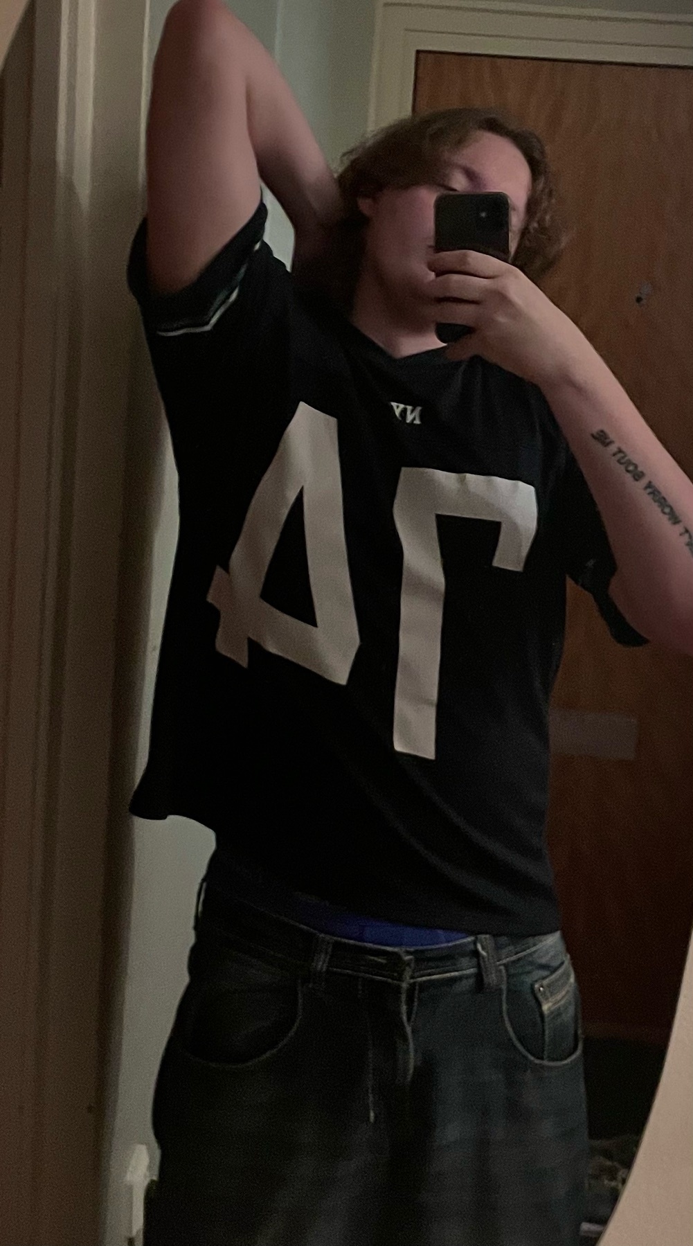 A man poses in front of a mirror, taking a selfie. He is shirtless and wearing jeans and a t-shirt.