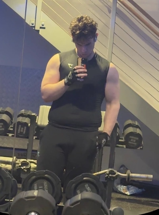A man in a black sleeveless shirt and black pants takes a mirror selfie in a gym. He is wearing black workout gloves and has curly brown hair. Dumbbells are visible in the foreground and background.