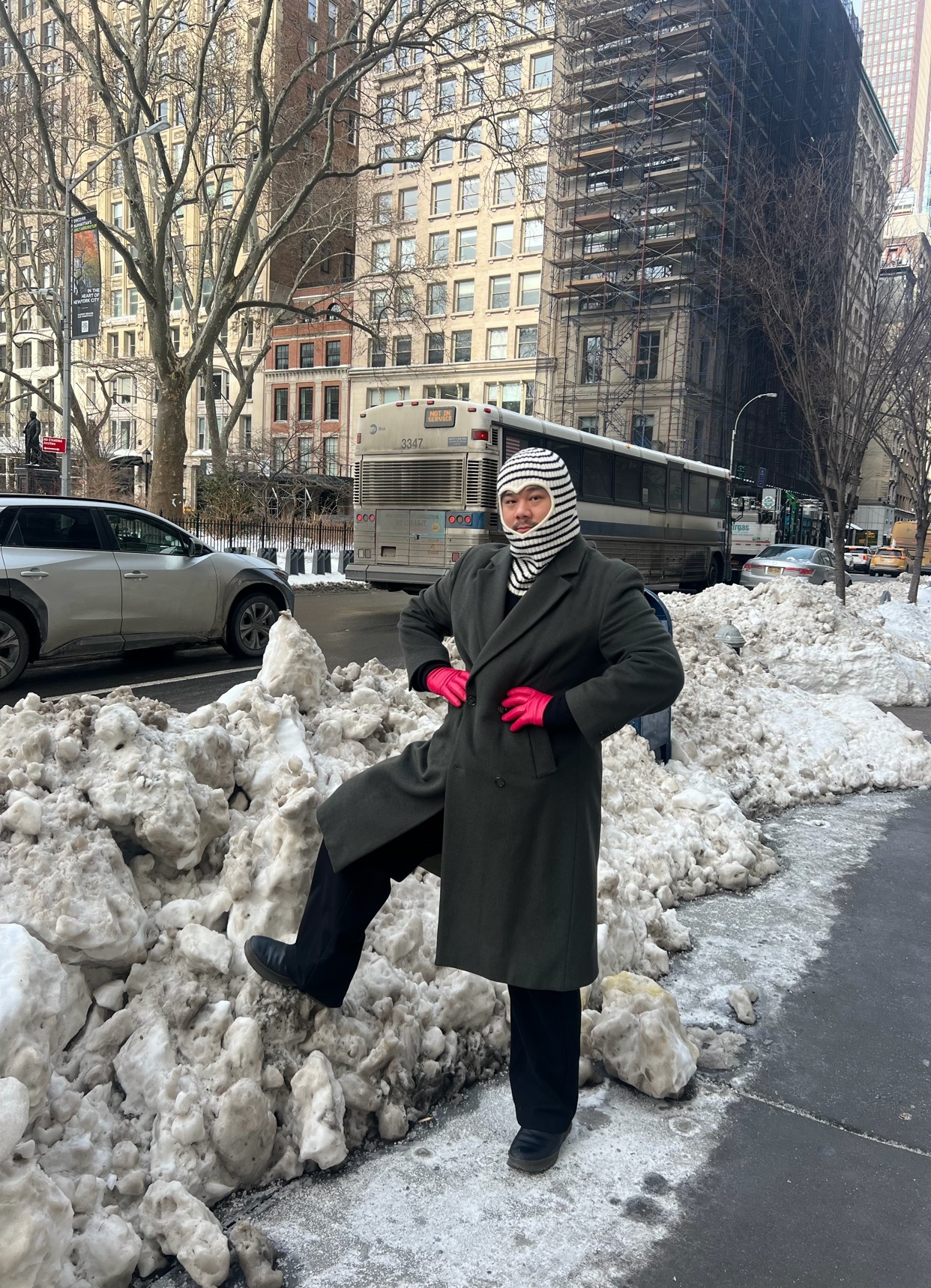 A man poses in a winter outfit next to a pile of snow in a city. He wears a balaclava, coat, and gloves.