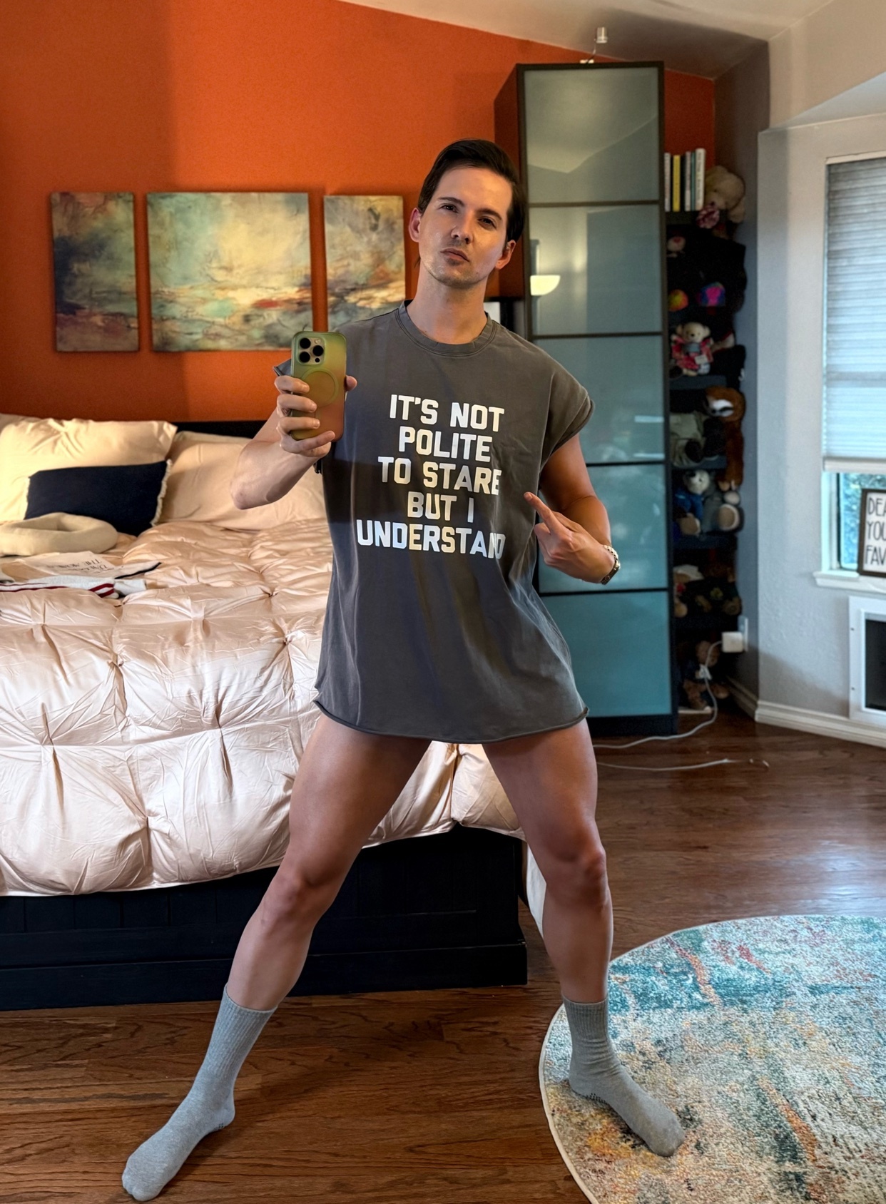 A man stands in a bedroom, wearing an oversized t-shirt with text that reads "IT'S NOT POLITE TO STARE BUT I UNDERSTAND." He is holding a phone and a drink, striking a confident pose.