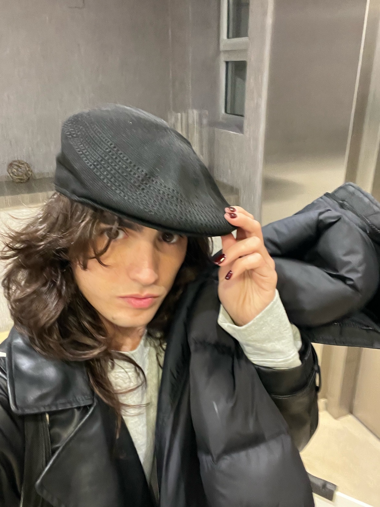 A man poses for a selfie, wearing a black leather jacket and a hat. The photo has a modern aesthetic.