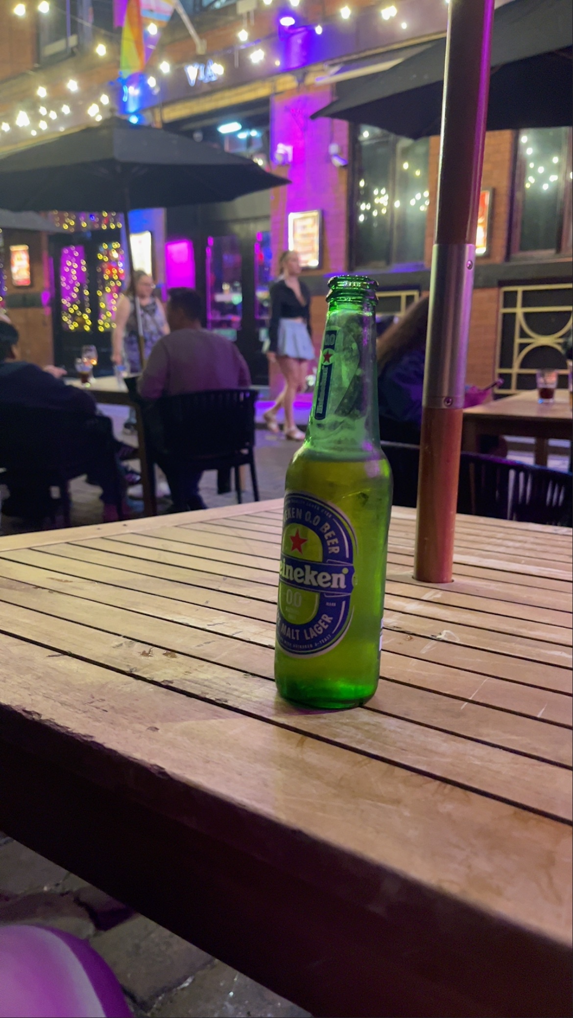 A Heineken beer bottle sits on a wooden table outside a bar or restaurant at night. In the background, people are visible walking and sitting, with colorful lights illuminating the street.