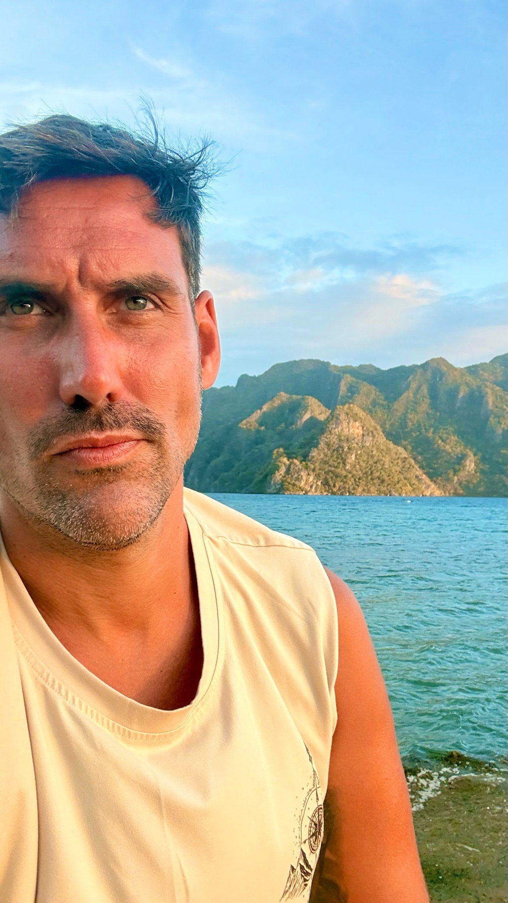 A man with stubble and a mustache takes a selfie by the ocean with mountains in the background under a cloudy sky.
