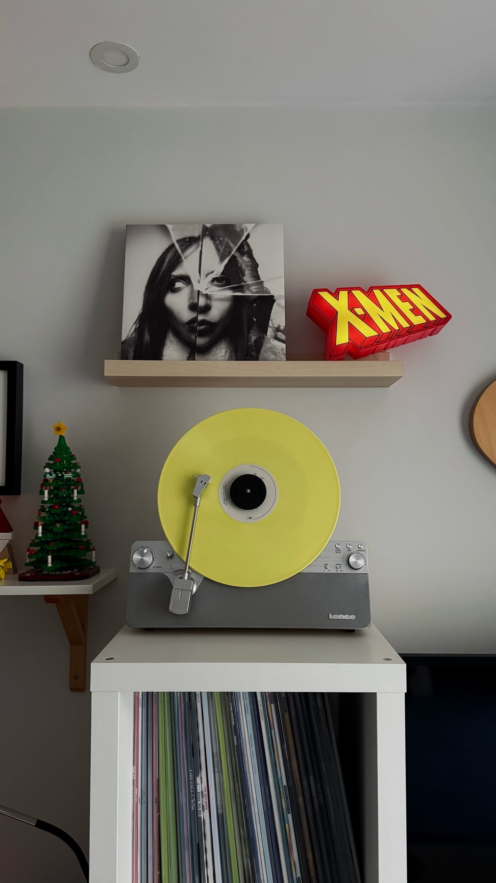 A record player with a yellow vinyl record on top of a white shelf. The shelf also holds an album cover and an X-Men sign.
