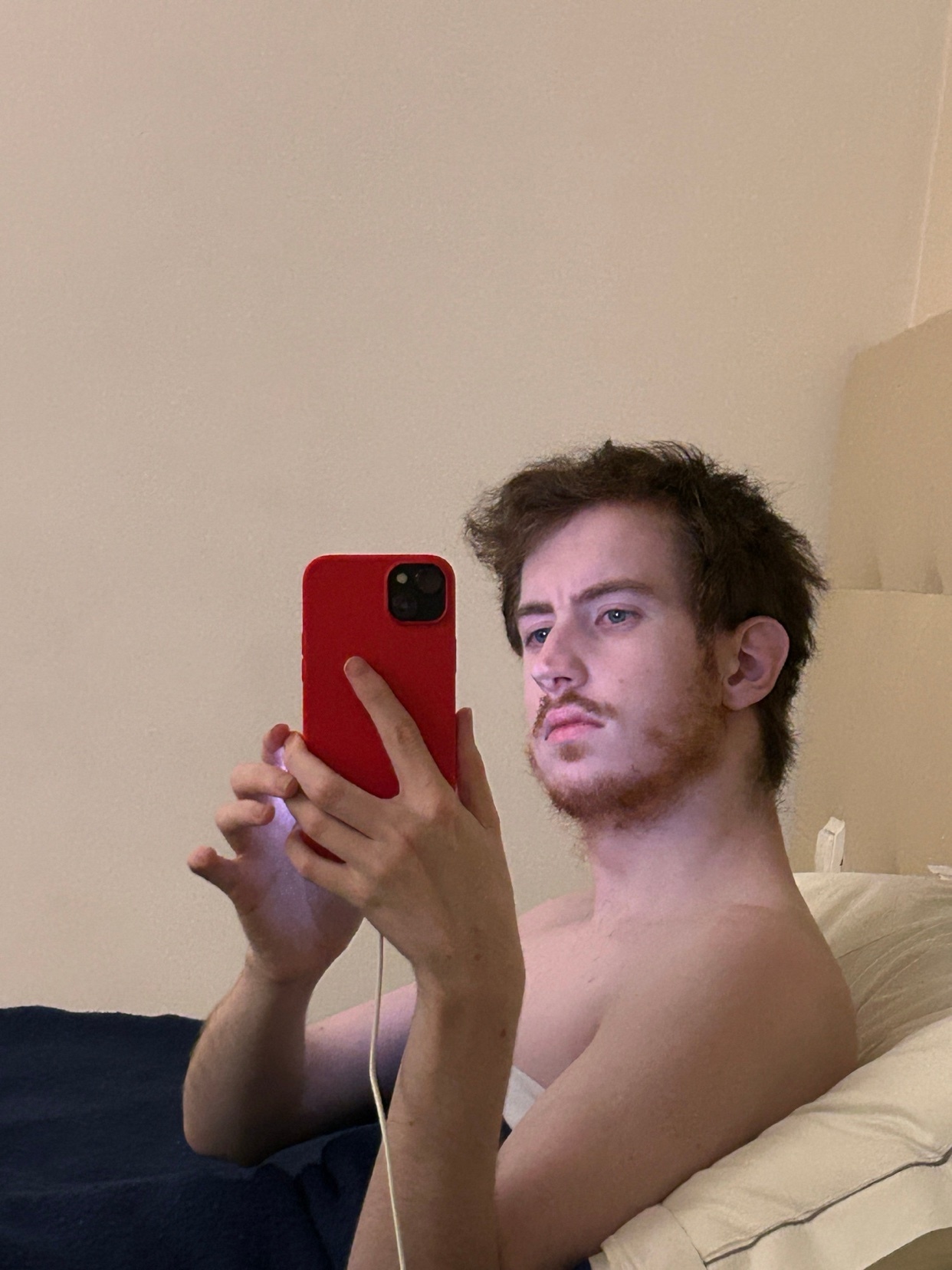 A man takes a selfie in his bedroom, shirtless. He has dark hair and stubble.