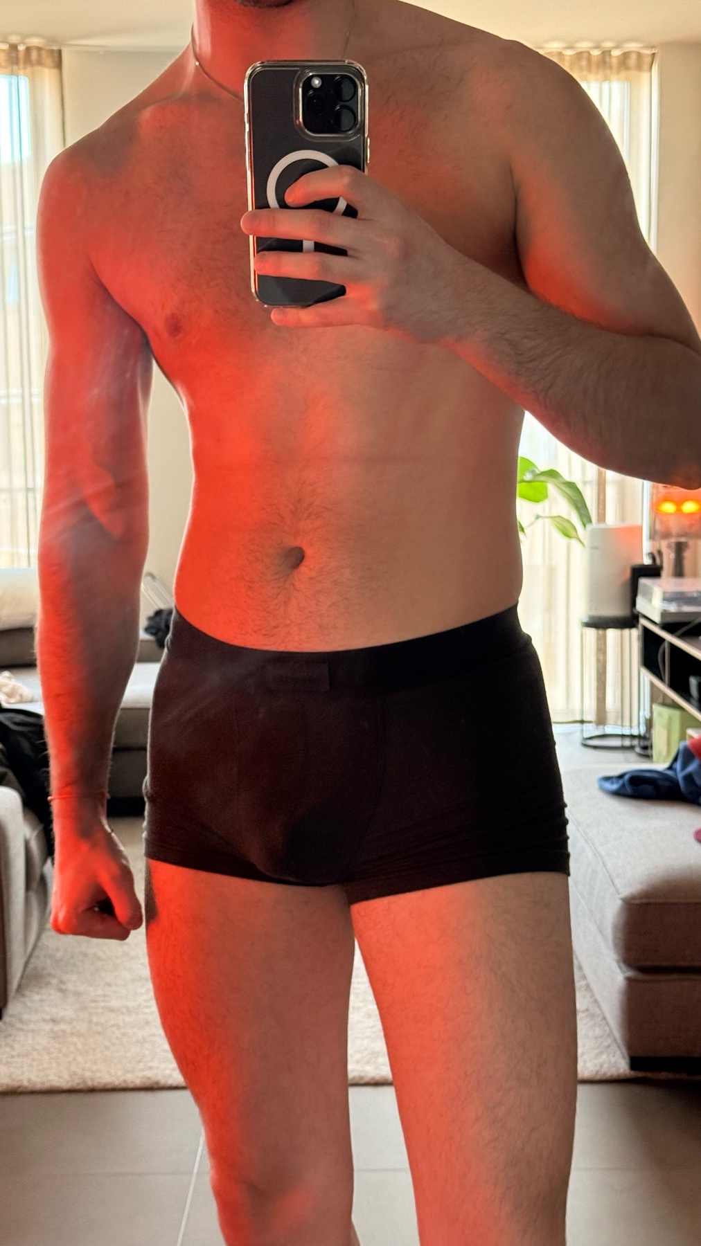 A man in black underwear takes a mirror selfie in a room with red lighting. He is flexing his bicep and his torso is bare.