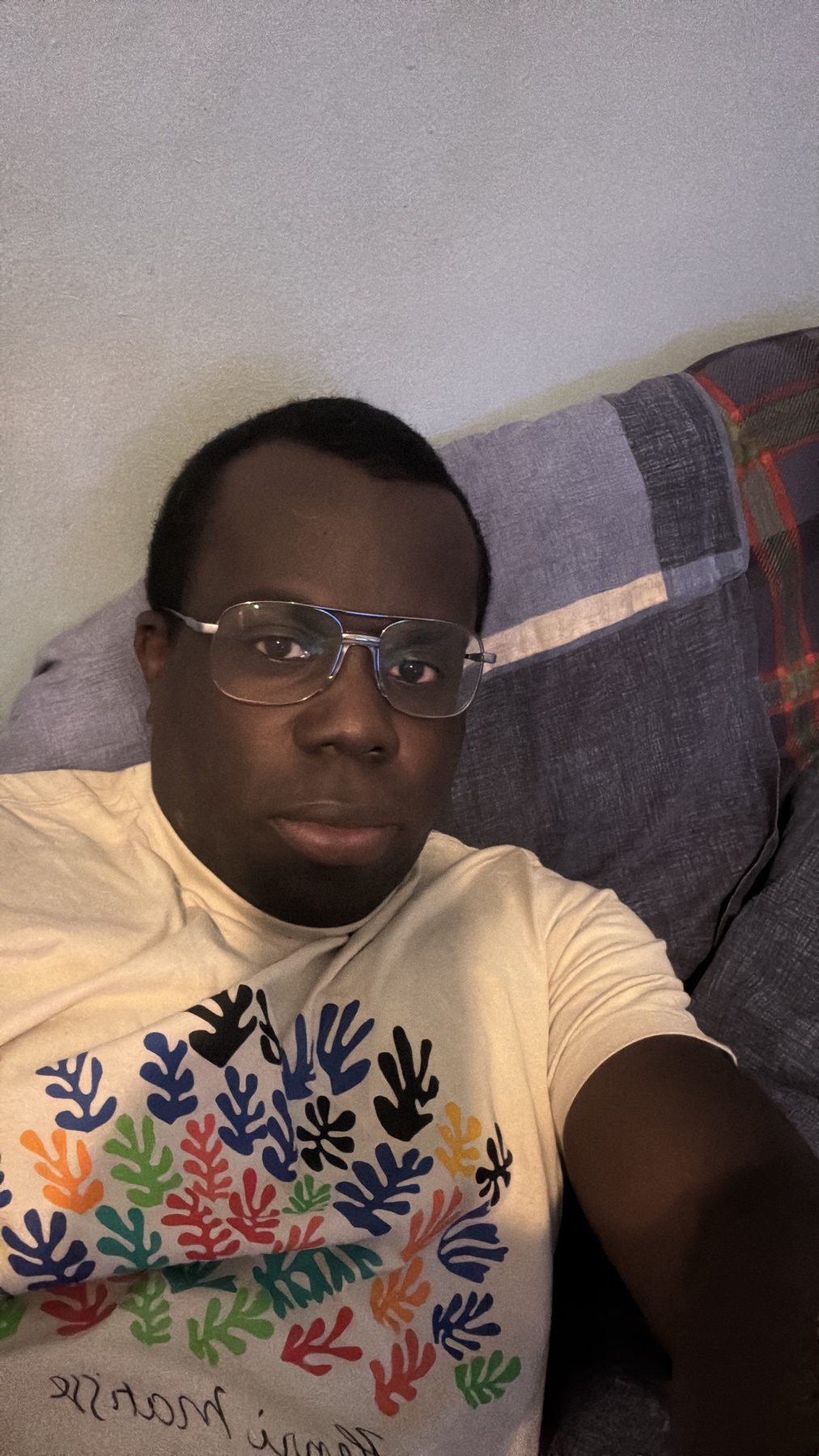 A man wearing glasses and a t-shirt poses for a selfie. The image is taken indoors.