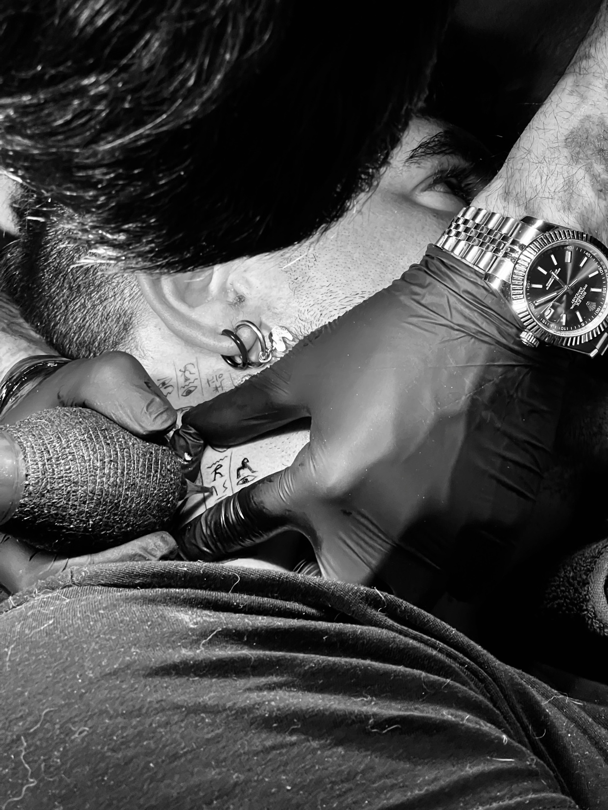 A close-up black and white photo of a man getting a tattoo on his neck. The tattoo artist's gloved hand is visible, holding the tattoo machine.