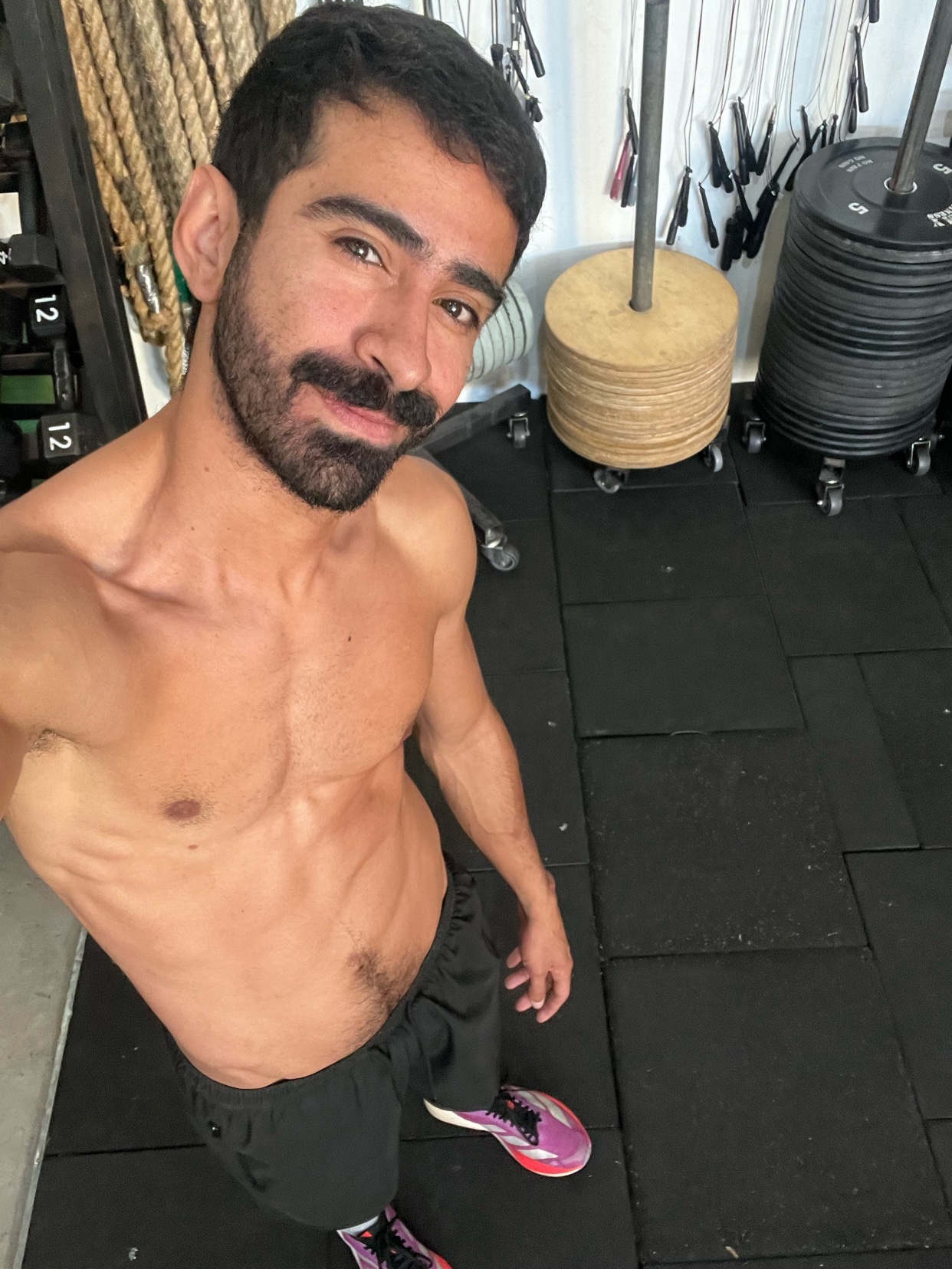 A man with a mustache and beard poses for a selfie in a gym. He is shirtless and wearing athletic shorts and sneakers.