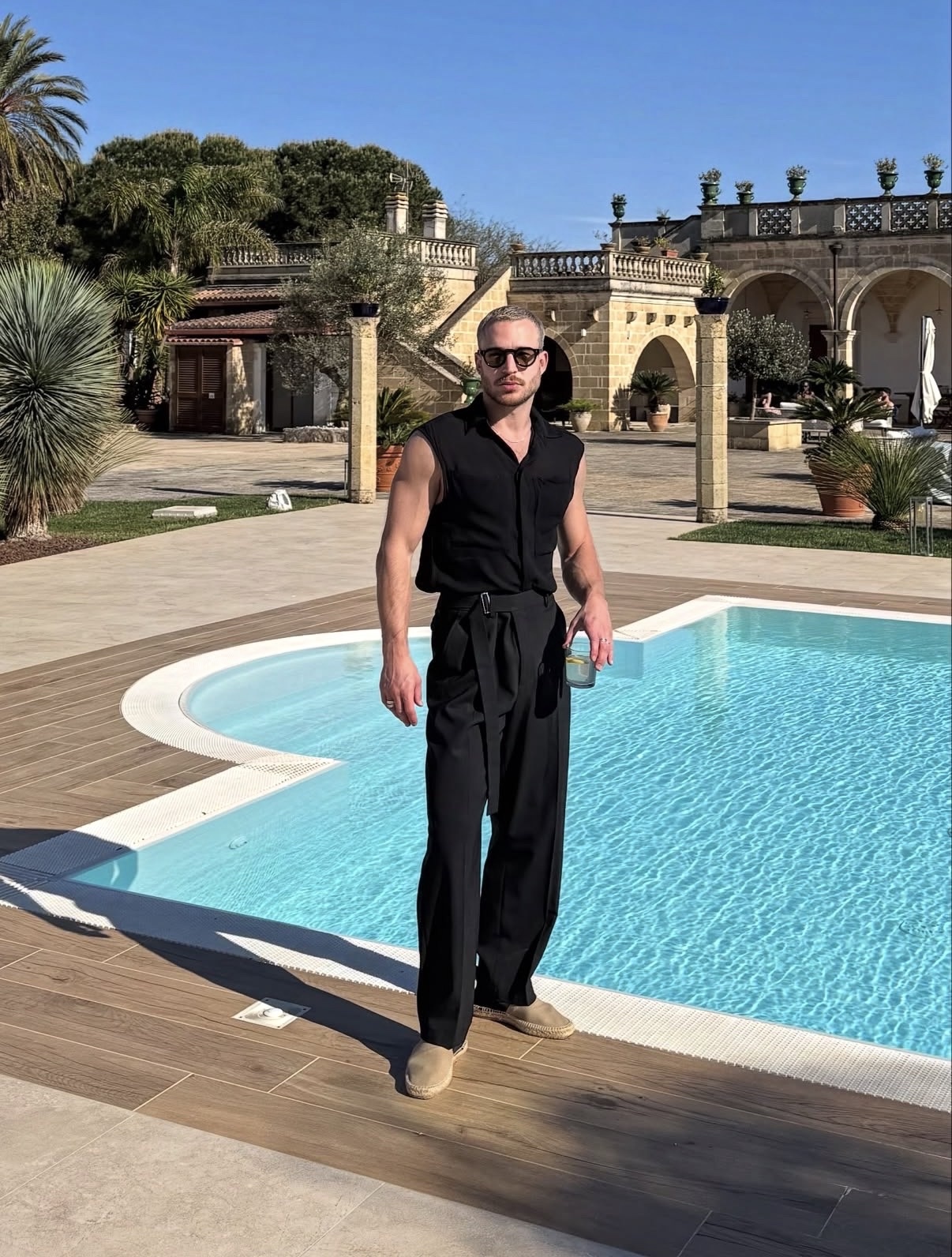 A man in a sleeveless black shirt and black pants poses poolside with sunglasses on. He holds a drink in his hand, and the background features a large building with arched windows and lush greenery.