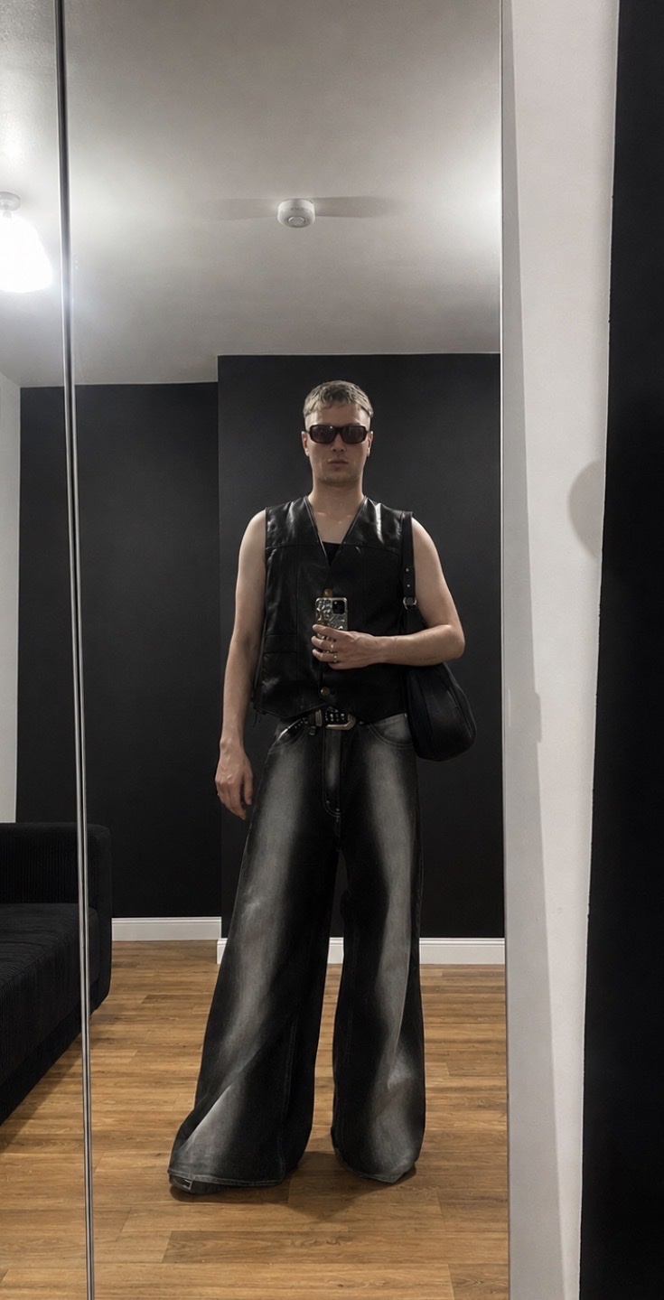 A man stands in front of a mirror, wearing a black vest, oversized leather pants, and sunglasses. He holds a phone in his hand and carries a black bag over his shoulder.