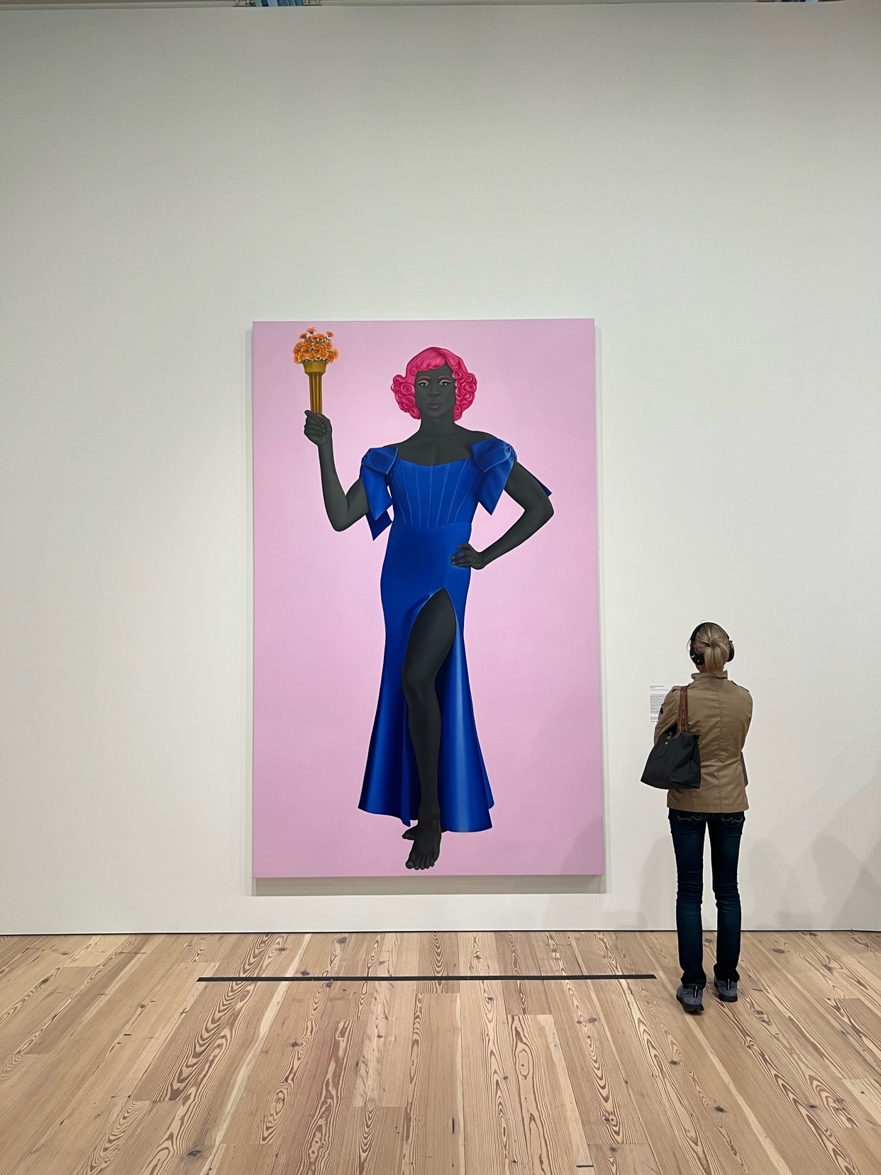 A woman stands in front of a large painting of a drag queen. The painting is in a gallery setting.