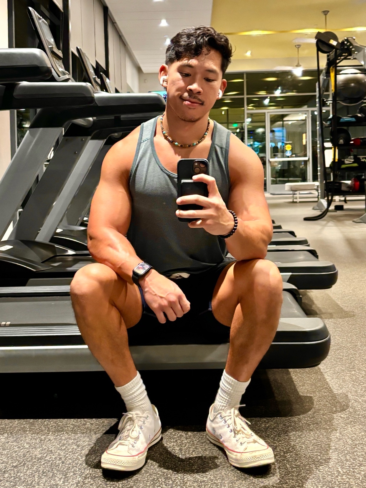 A man takes a selfie at the gym. He is muscular and wearing a tank top and shorts.
