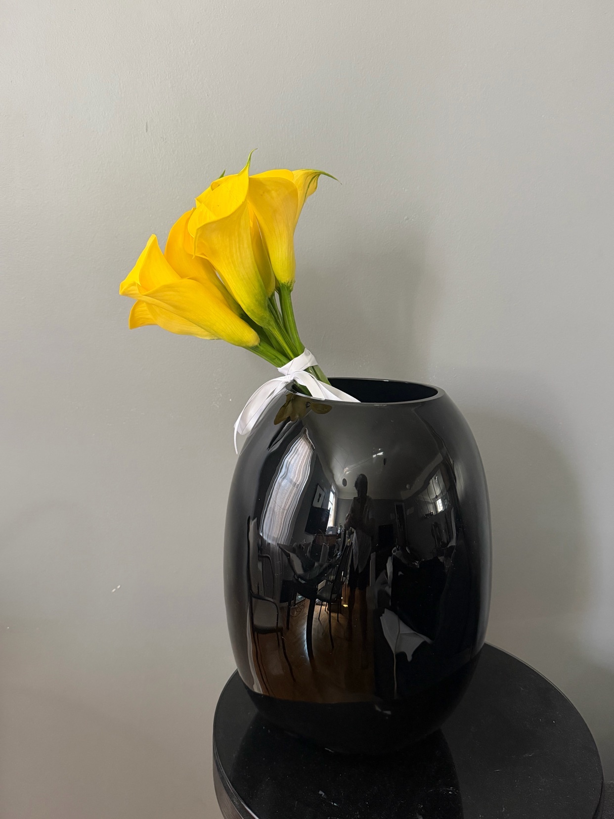A bouquet of bright yellow calla lilies is tied with a white ribbon and placed in a glossy black vase. The vase sits on a dark, reflective surface against a neutral gray wall.