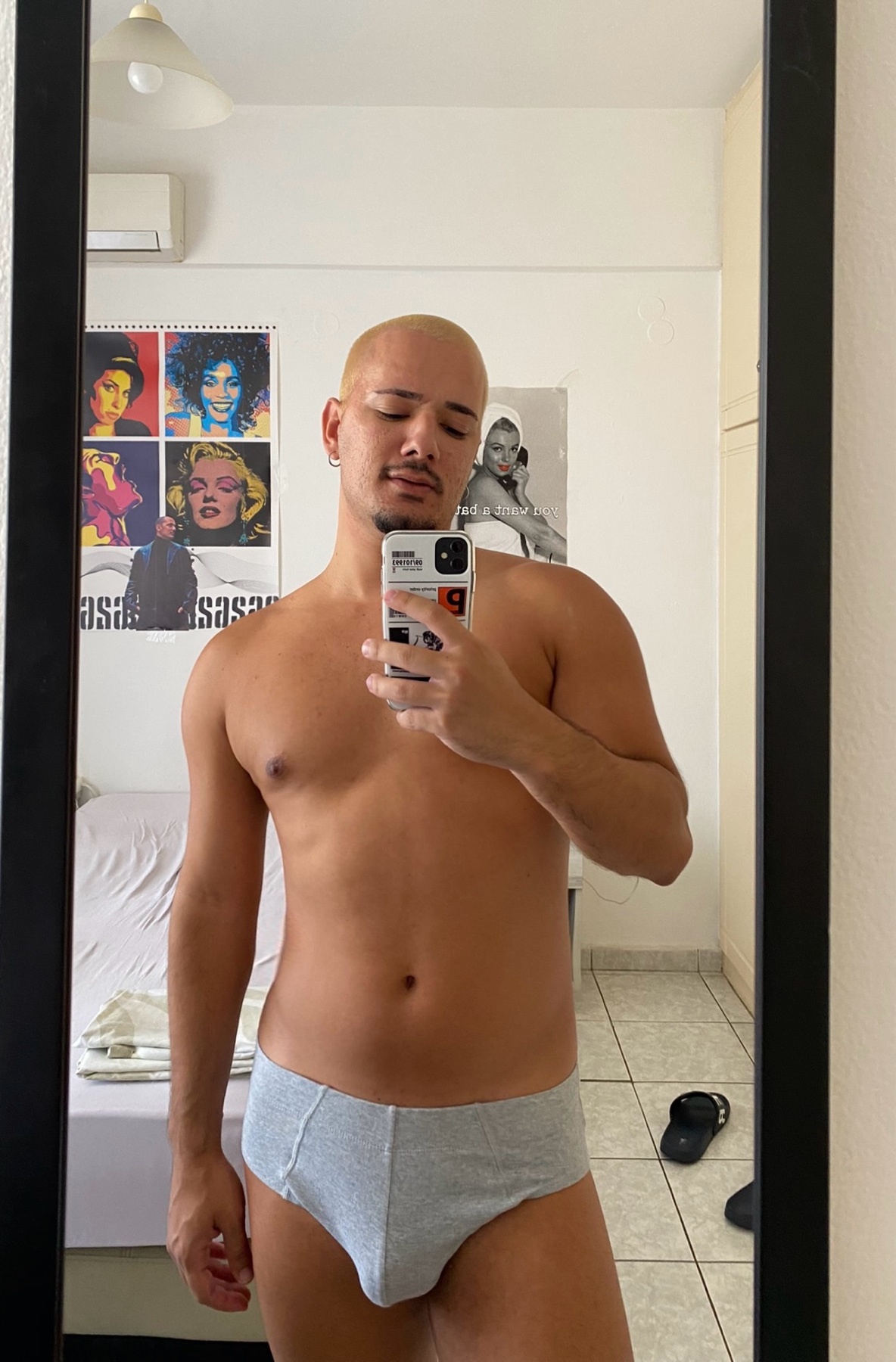A man takes a mirror selfie, shirtless and wearing underwear. He has a bald head and is holding a phone.