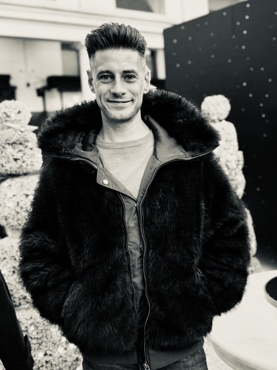 A man poses in a black fur coat. The black and white photo highlights the texture of the coat and the man's stylish look.