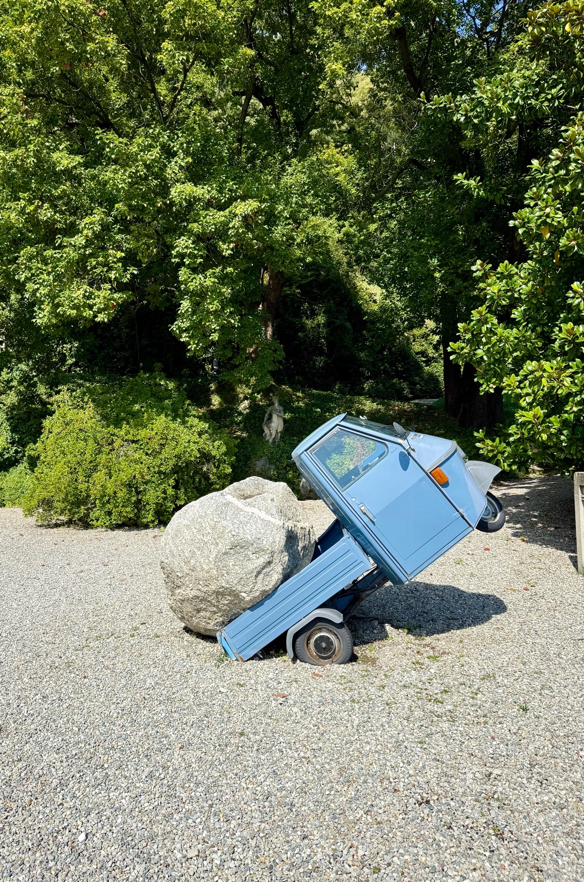 A light blue, three-wheeled vehicle is tilted precariously, with its cargo bed resting on a large, rough boulder. The scene is set outdoors with lush green trees and gravel on the ground.