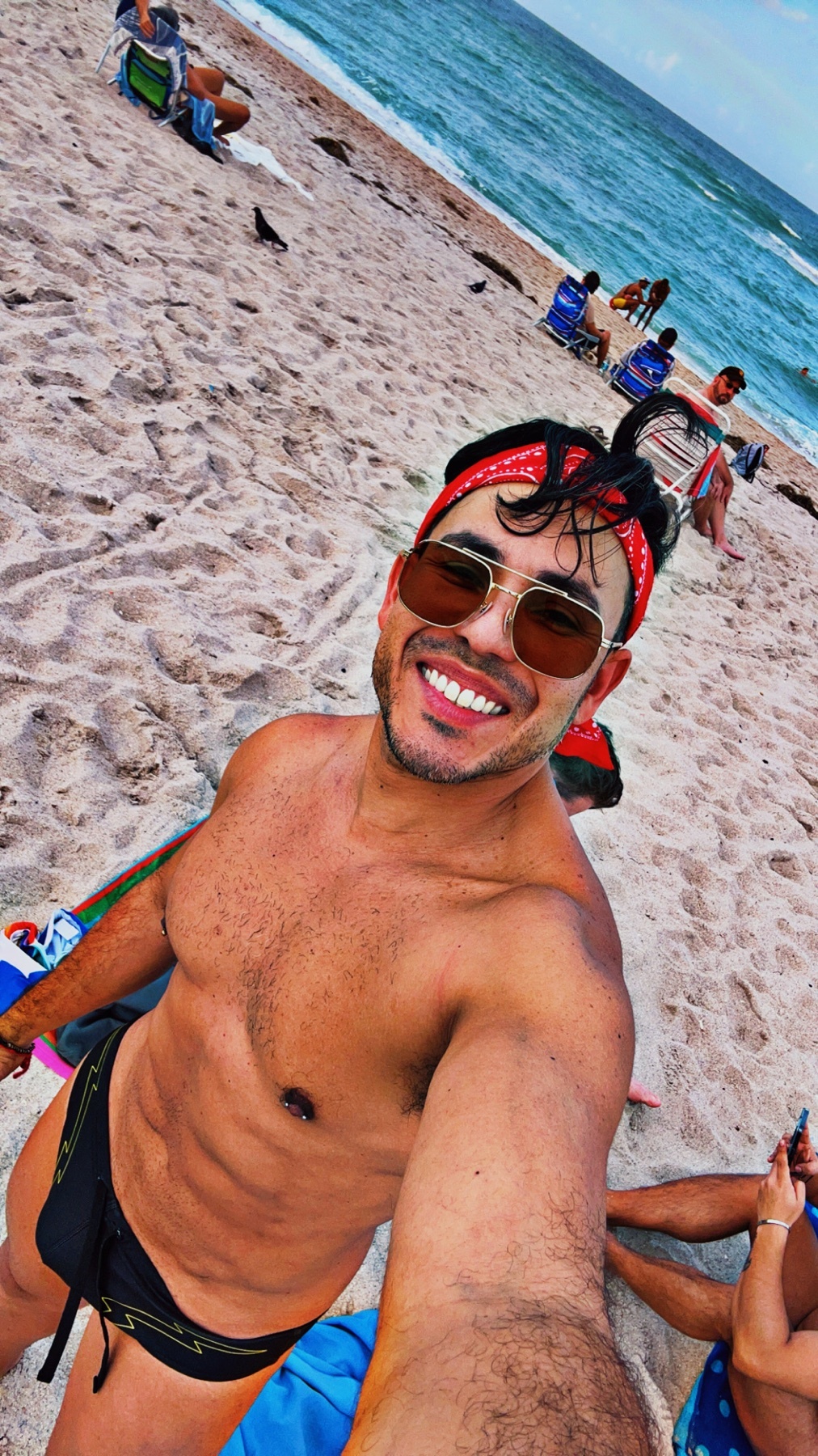 A man with a bandana and sunglasses smiles at the camera while taking a selfie on a sunny beach. He is wearing black swimwear and appears to have a muscular physique.