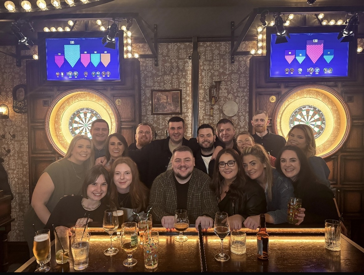 A group of friends poses for a photo behind a bar with darts boards and screens in the background.