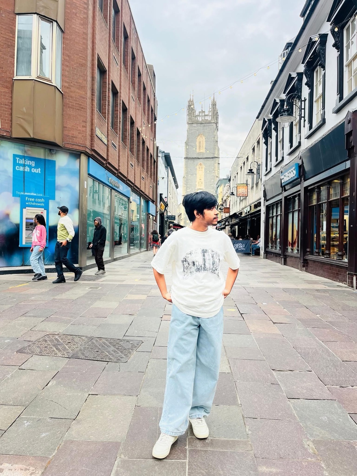 A man poses on a cobblestone street in front of a historic building. He wears a t-shirt, light blue jeans, and white sneakers.