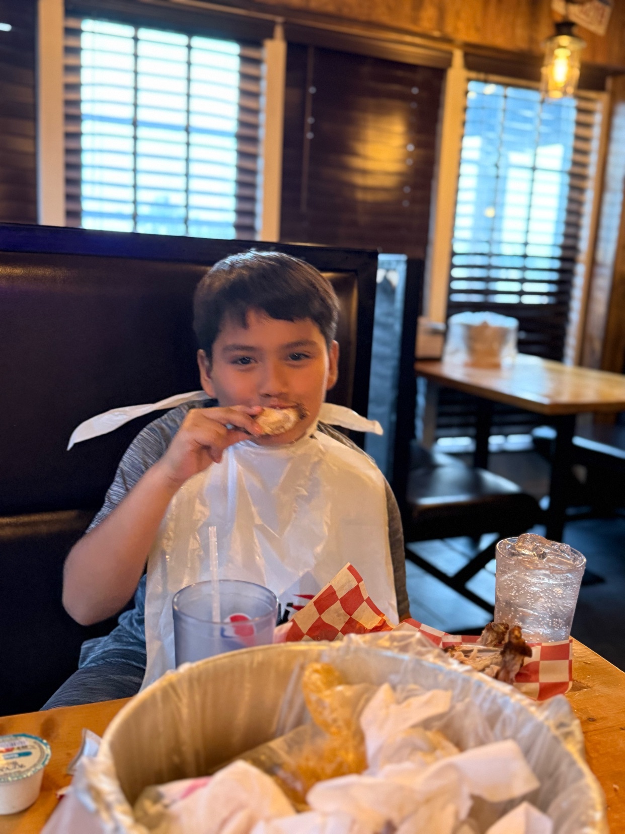 A young boy wearing a bib is sitting at a restaurant table and eating a piece of food. There is a basket of food and drinks on the table.