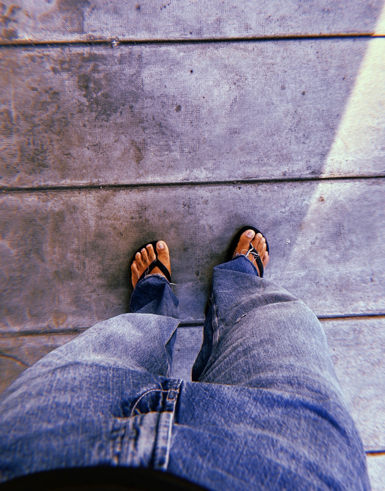 A first-person view of a person wearing blue jeans and flip-flops standing on a wooden deck.