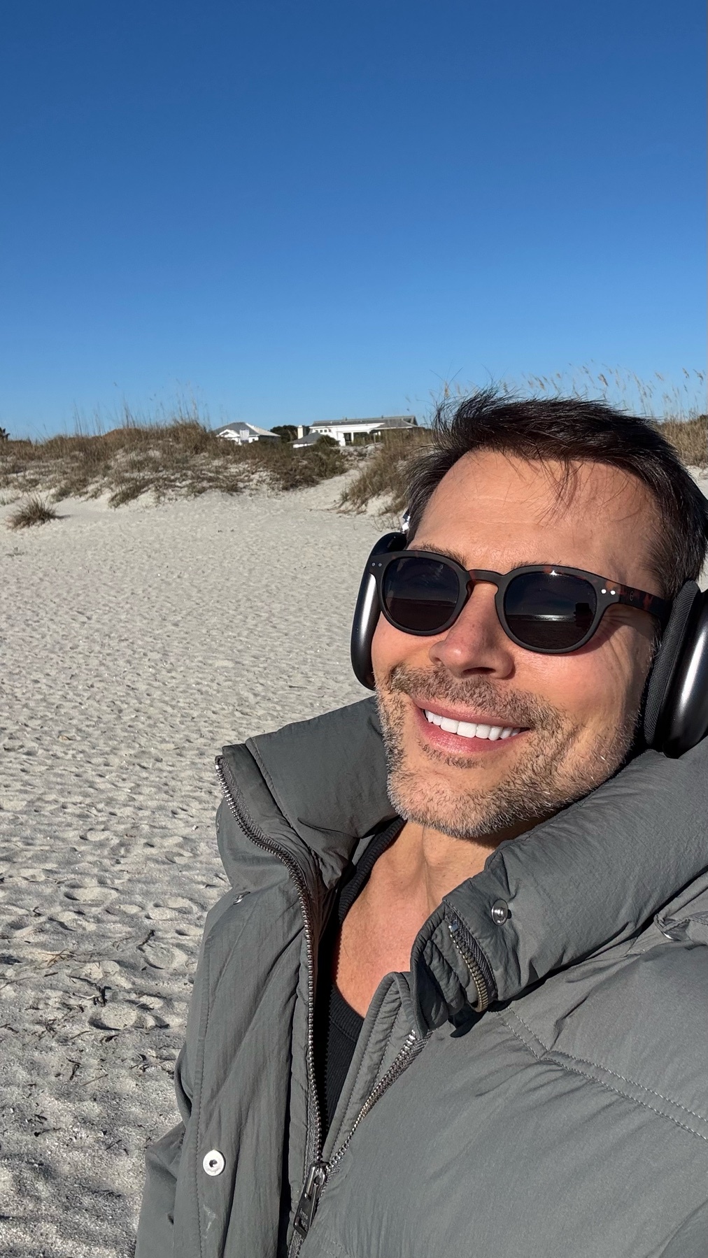A man smiles while wearing sunglasses and headphones on a sunny beach day. He is wearing a grey jacket.